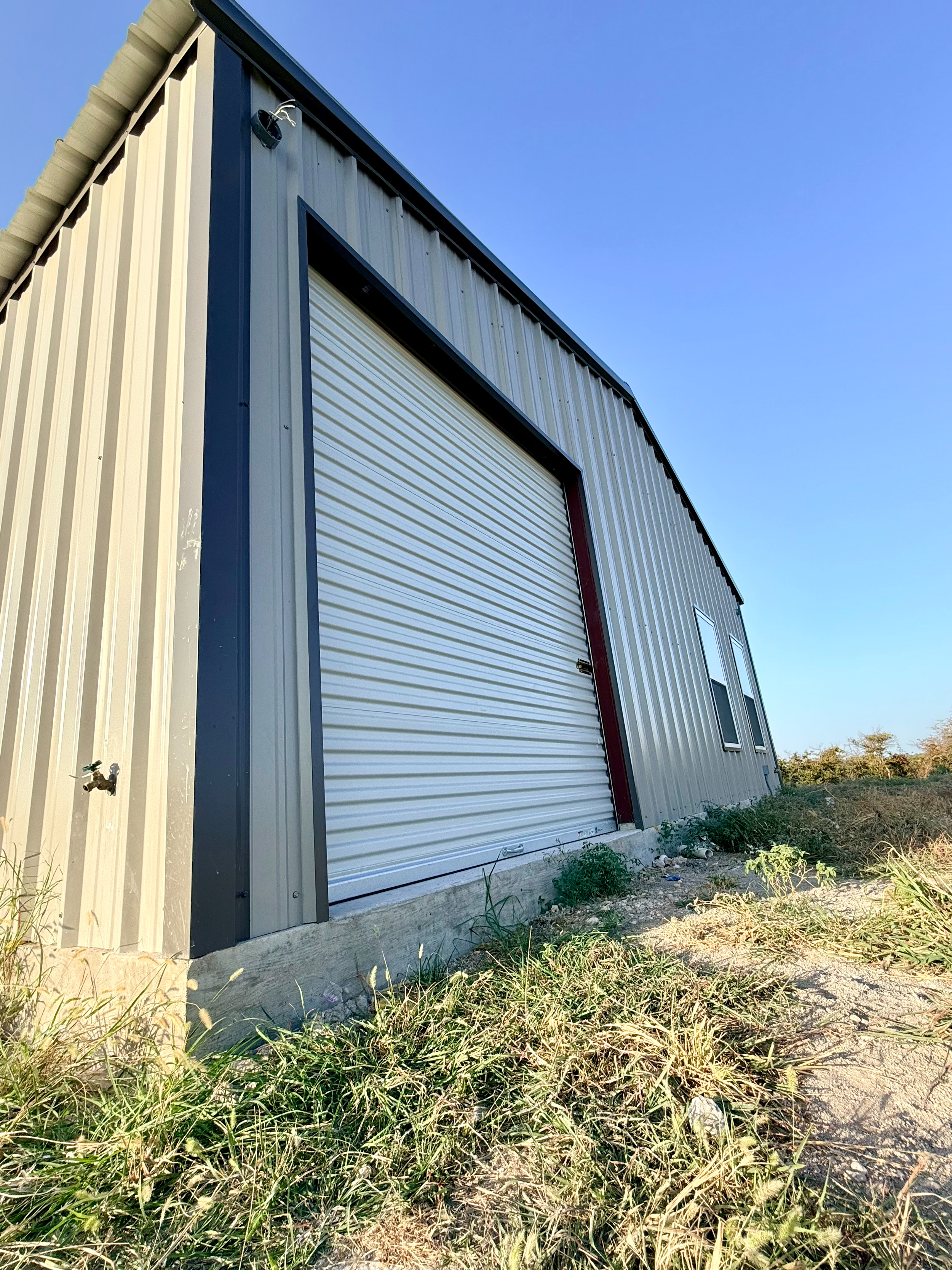 A metal building with off-white siding, black trim, and a rolled-down white garage door, situated on a concrete foundation with overgrown grass and weeds in the foreground, under a clear blue sky.