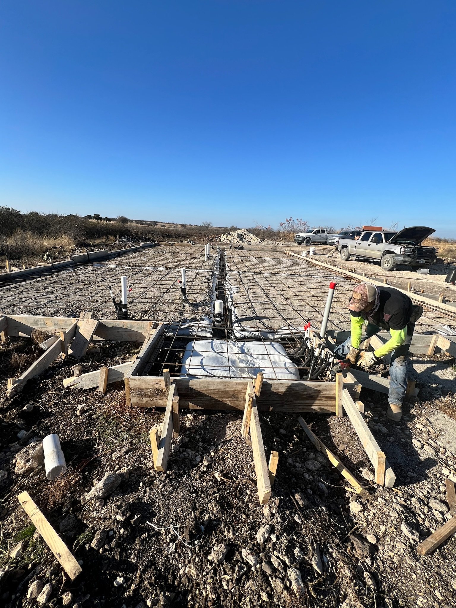 Construction workers install rebar for a concrete foundation on an outdoor construction site with trucks parked nearby and open land under a clear blue sky.