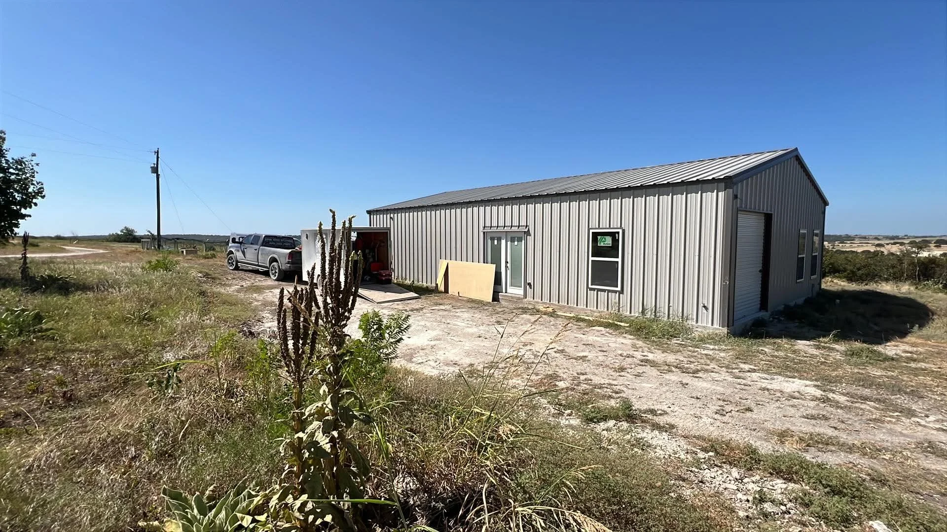 A metal building with a sloped roof and a garage door in a rural area under a clear blue sky. A pickup truck is parked nearby, and there are some plants and dry grass in the foreground.