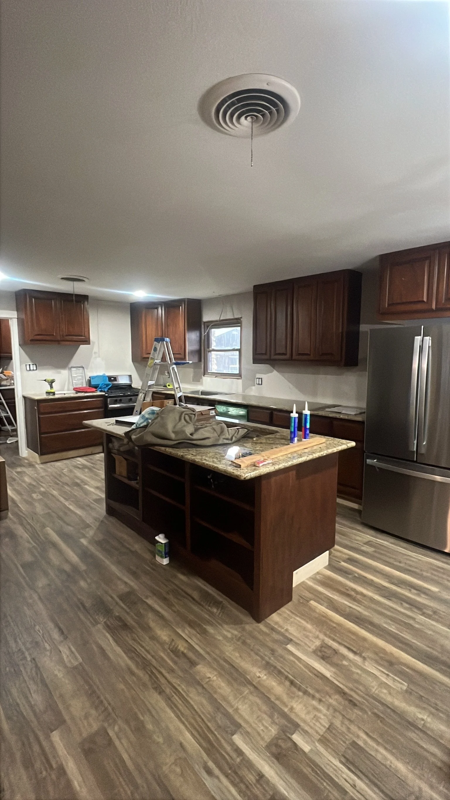 Modern kitchen with wooden cabinets, a granite island, stainless steel refrigerator, and unfinished ceiling with a smoke detector. There are tools and supplies on the island, a ladder, and construction materials around.