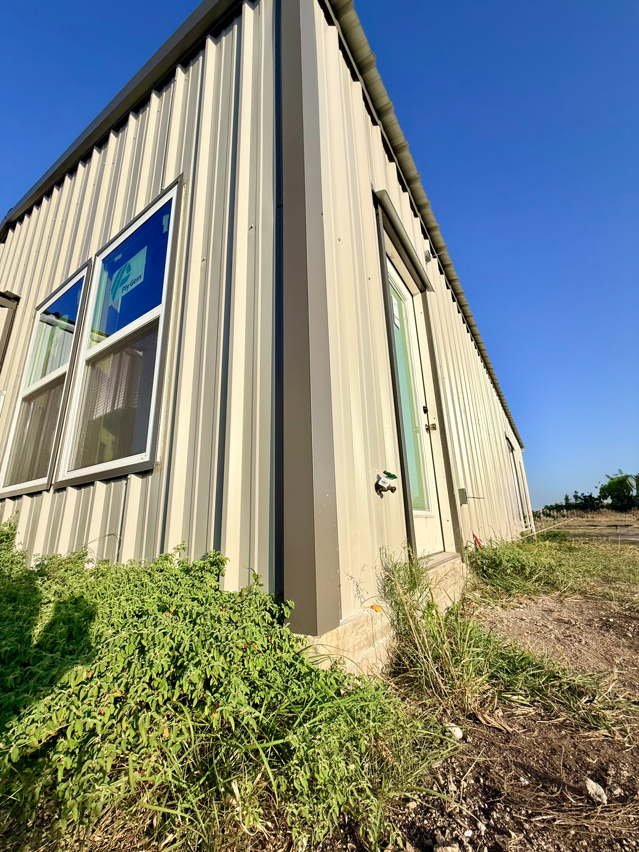 Side view of a metal building with vertical siding, multiple windows, and a door, situated on a concrete foundation with green bushes and grass in the foreground, under a clear blue sky.