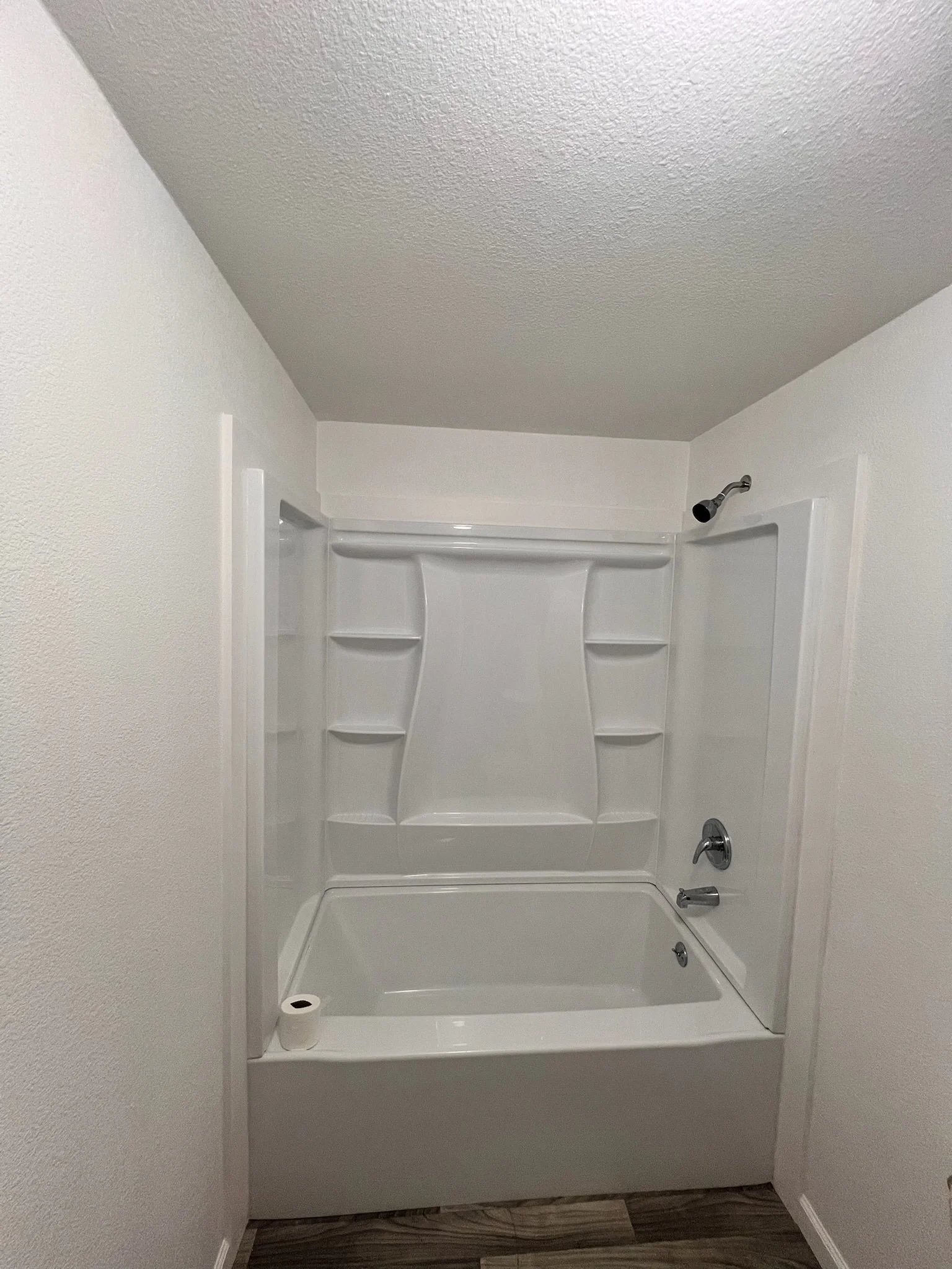 Bathroom shower with a white shower stall, built-in shelves, a shampoo holder, a showerhead, and a bathtub, with a toilet paper roll on the edge of the shower.