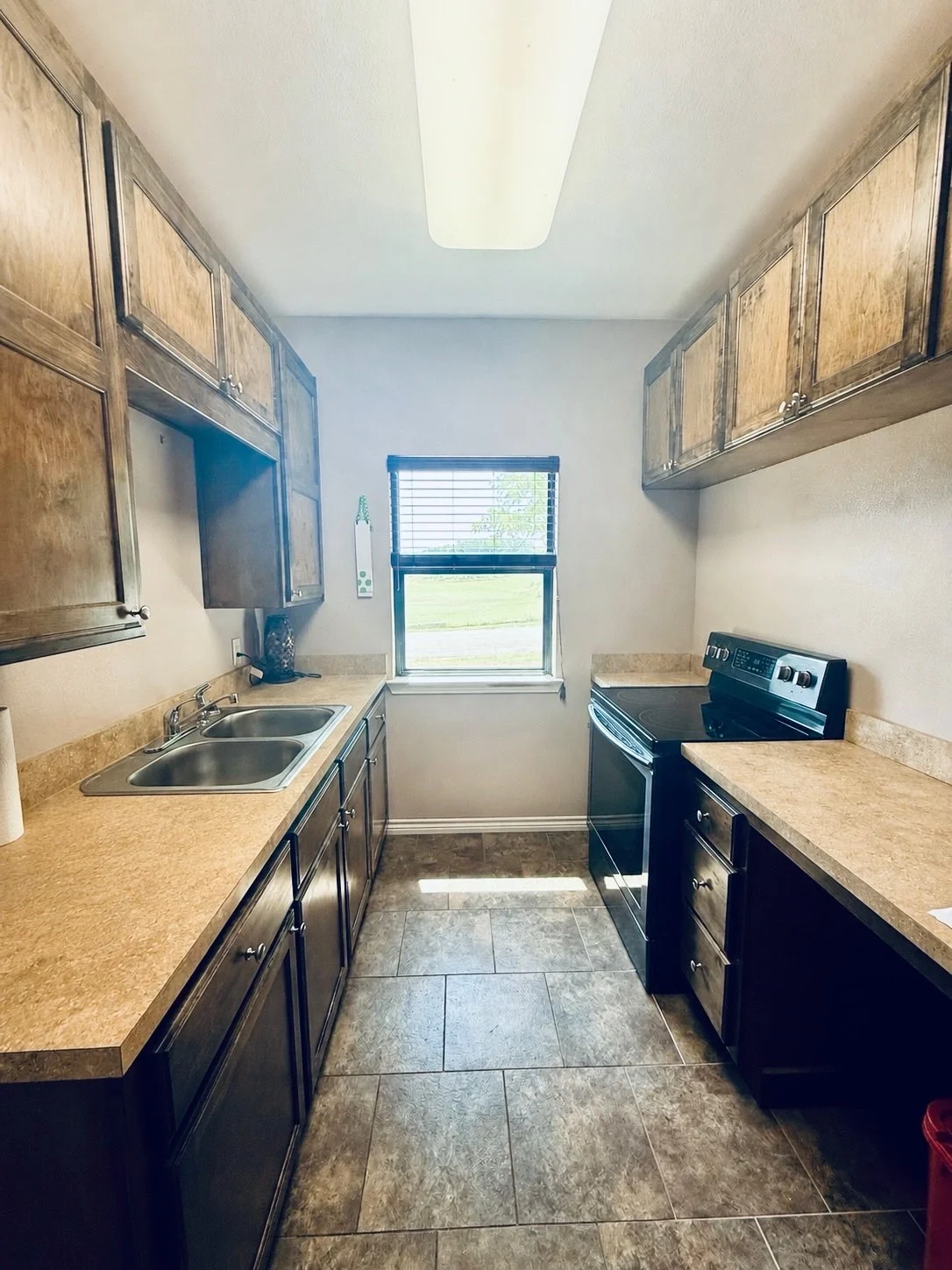 A small kitchen with dark wooden cabinets, beige countertops, a stainless steel double sink, a window with blinds, an electric stove, and tiled floor.
