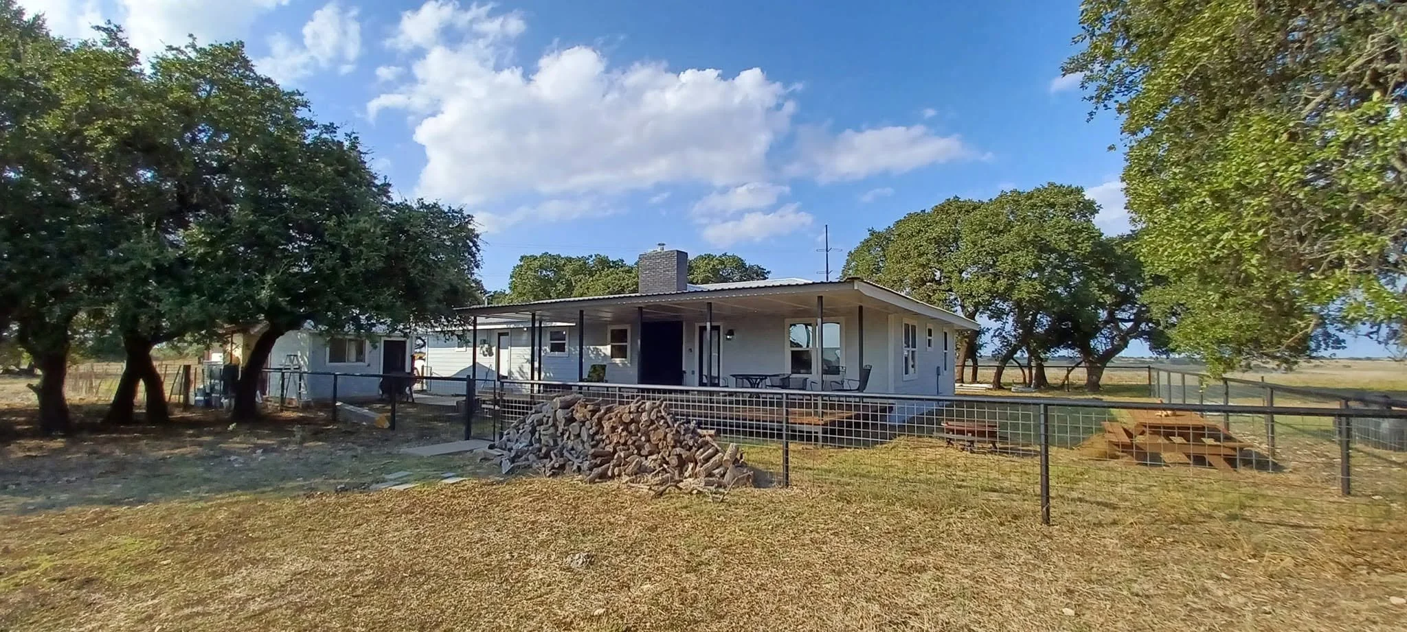 A single-story white house with a metal roof, surrounded by trees, and a fenced yard with a pile of firewood and stairs leading up to a porch.