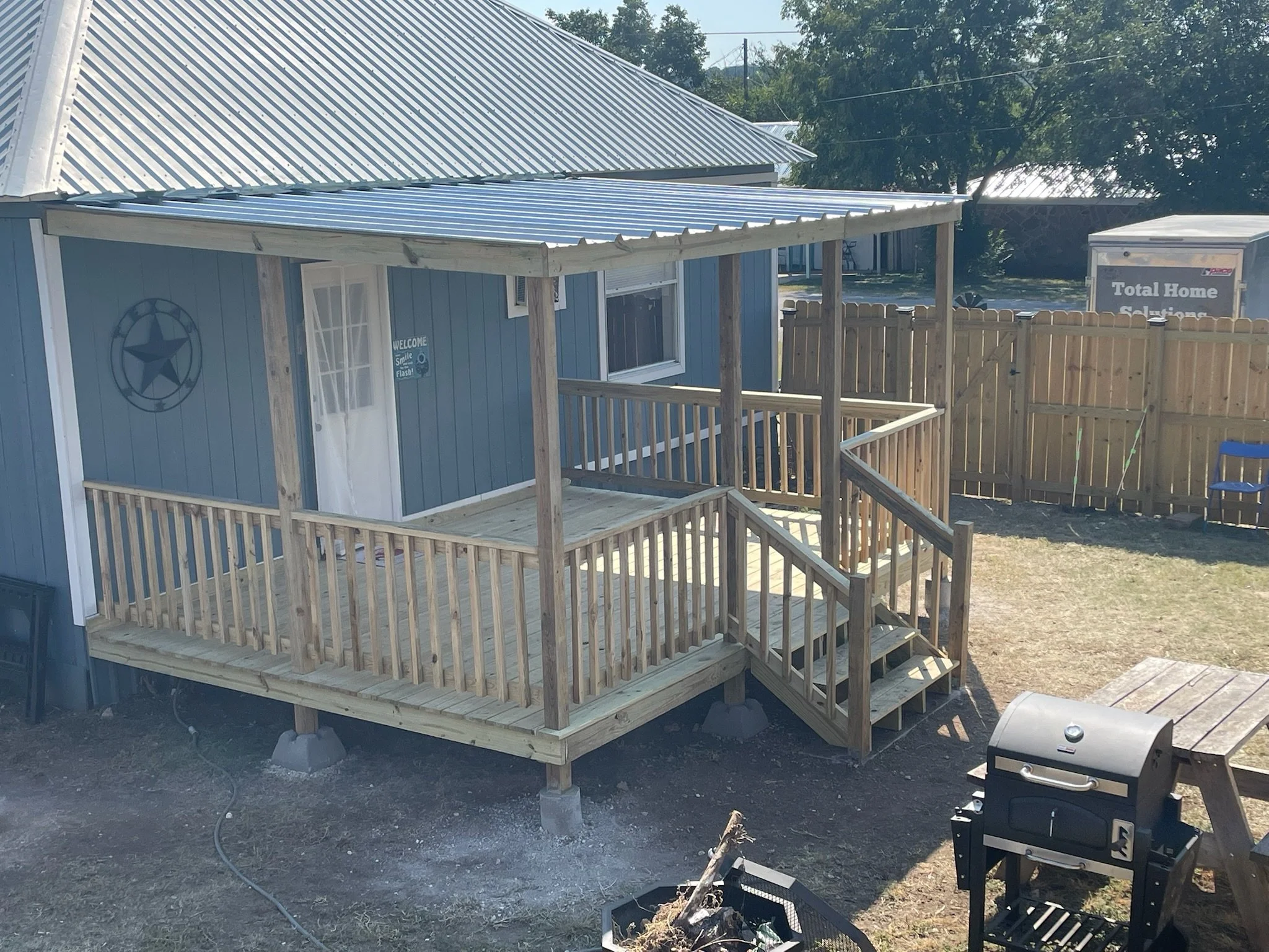 A small house with blue siding and a metal roof, surrounded by a wooden fence, with a deck and a Welcome sign near the door. There's a charcoal grill nearby and trees in the background.