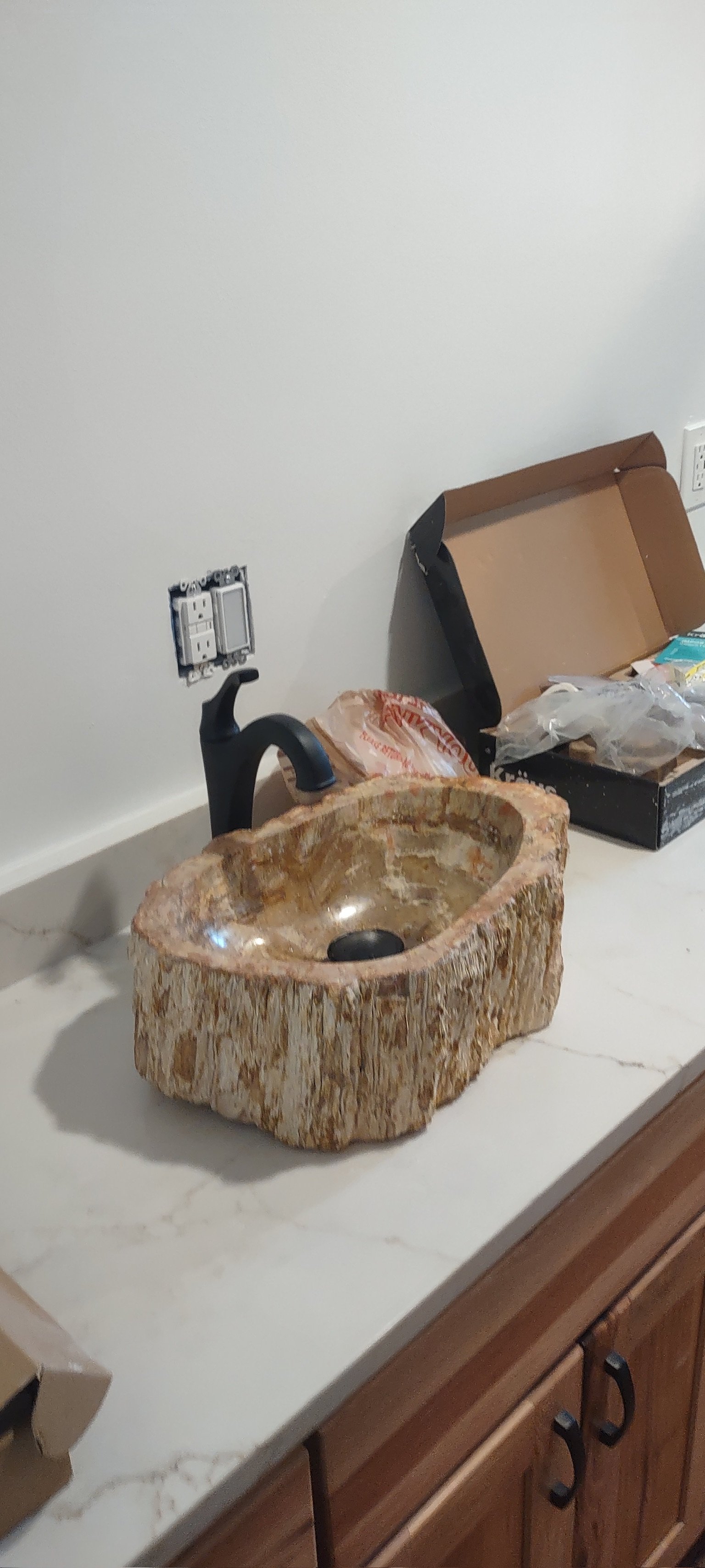 A rustic kitchen sink carved from a large piece of stone with a black faucet, placed on a white countertop. There is an open box and some plastic bags beside it, against a white wall with empty electrical outlets.