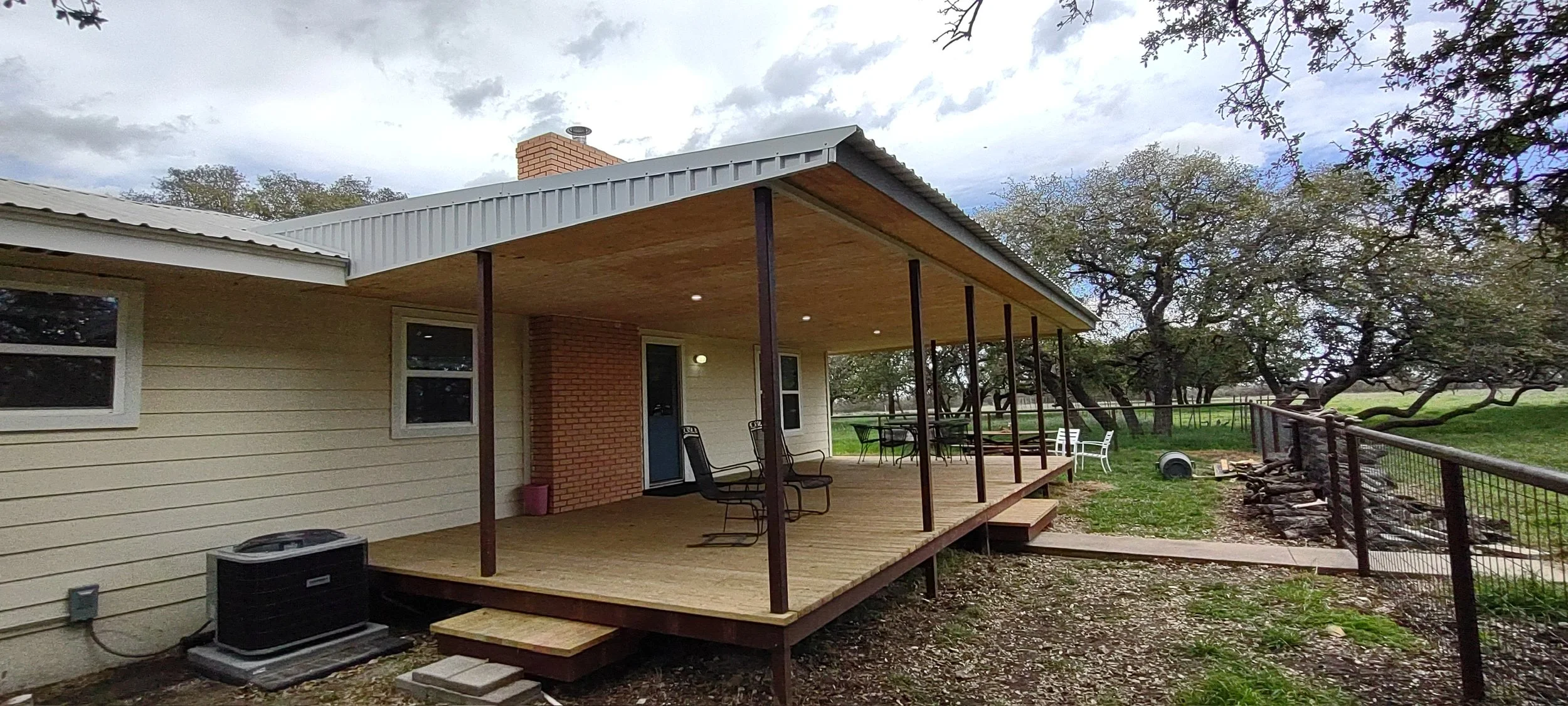 Back porch of a house with outdoor furniture, wooden deck, and trees in a grassy yard under a cloudy sky.