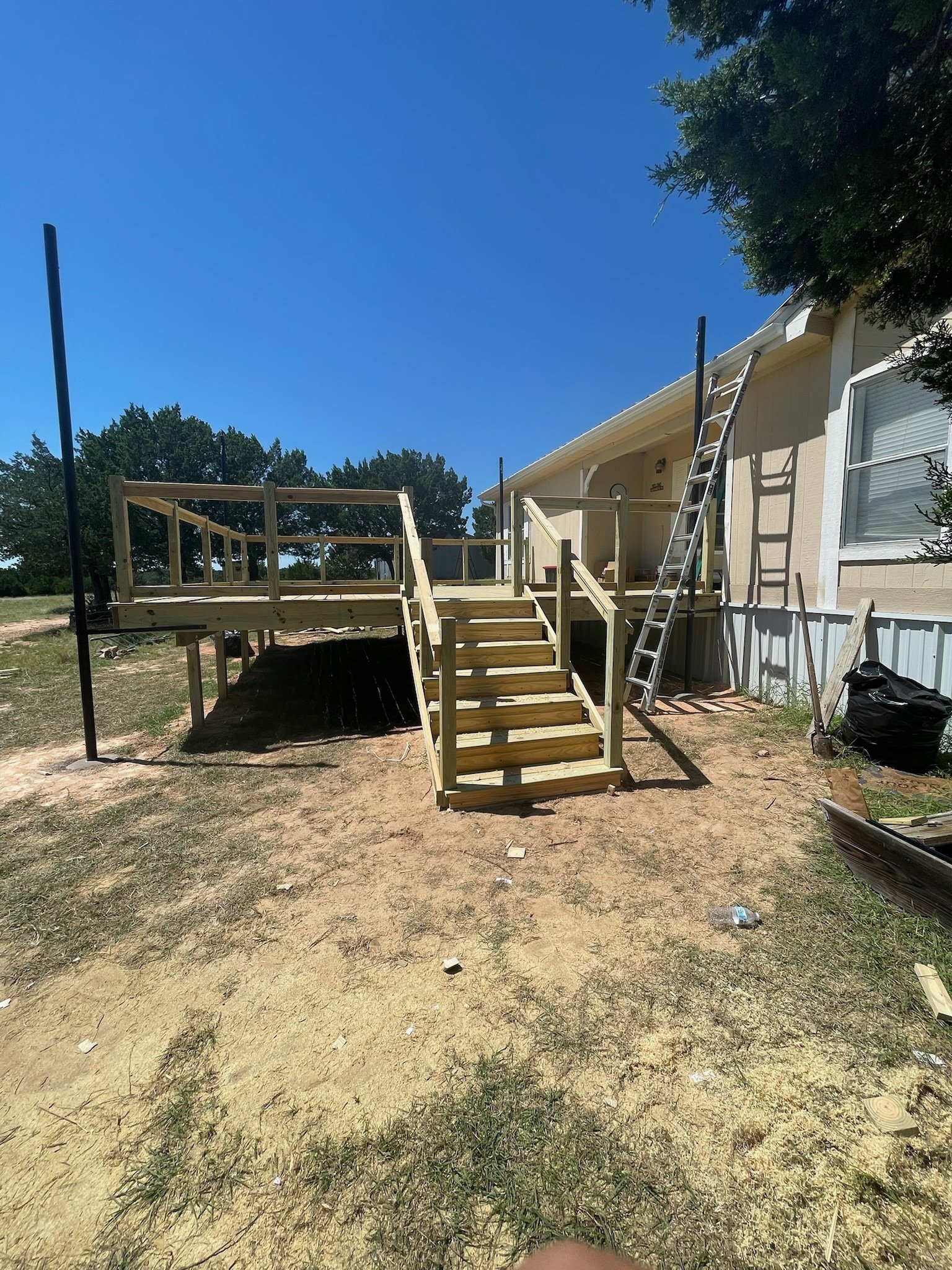 A house with a pale yellow exterior is under construction with a wooden deck and a ladder leaning to the side. The yard has a mix of bare soil and sparse grass, and there are trees and a clear blue sky in the background.