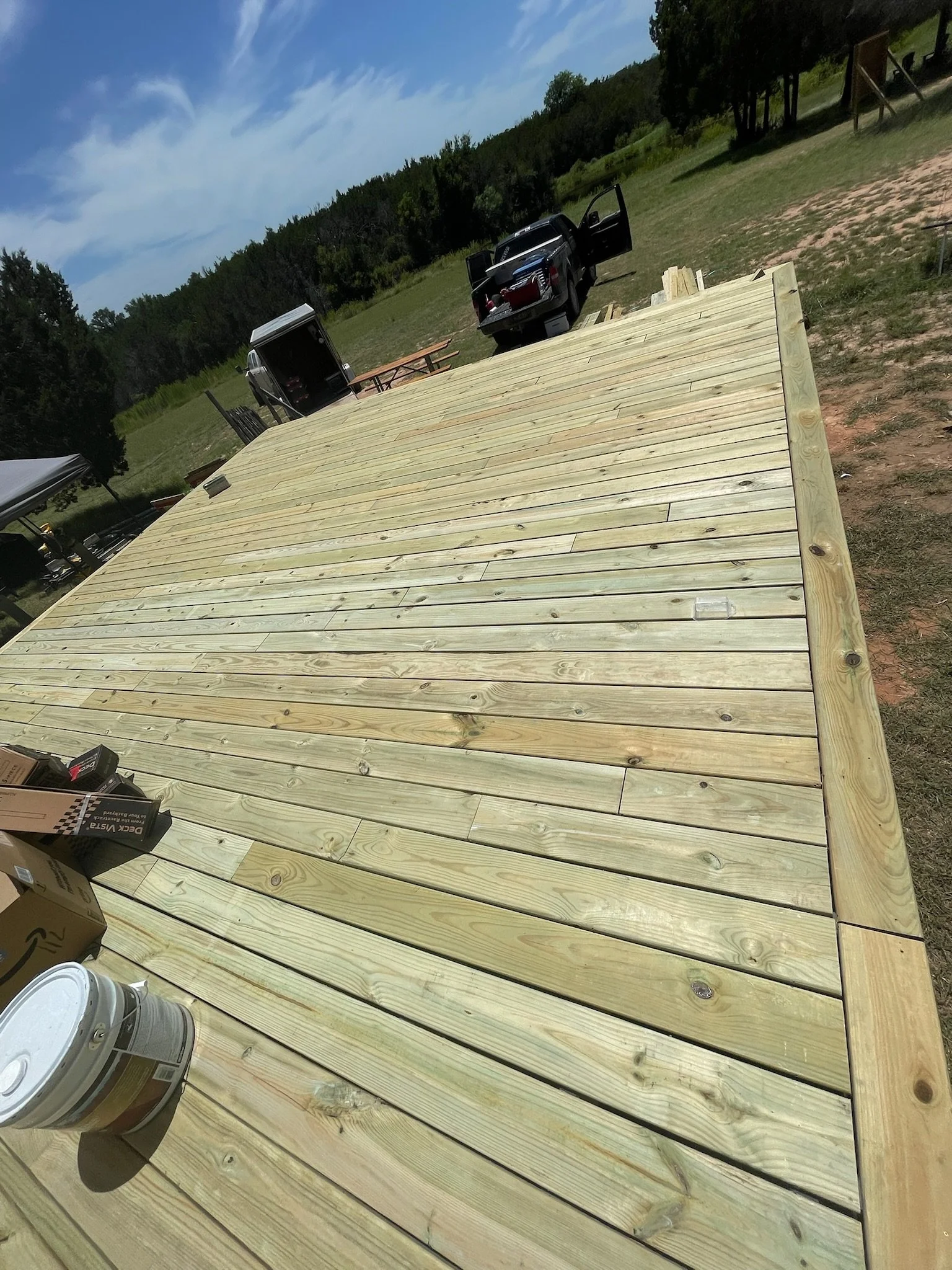 An outdoor scene of a large wooden deck under construction in a rural area with a grassy field, trees, a pickup truck, a trailer, and various construction supplies.