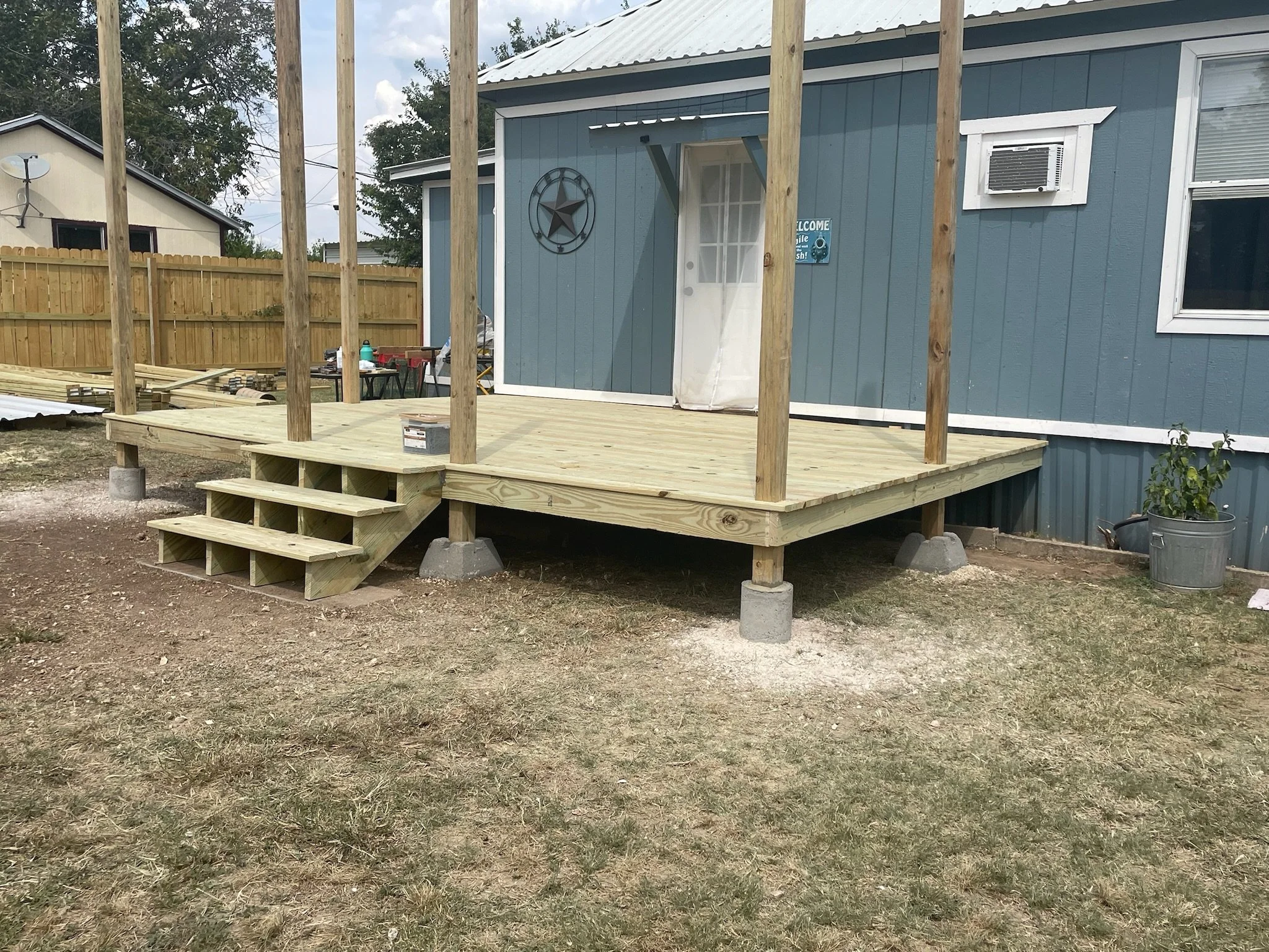 Backyard deck under construction with vertical support beams, steps, and a blue house in the background.
