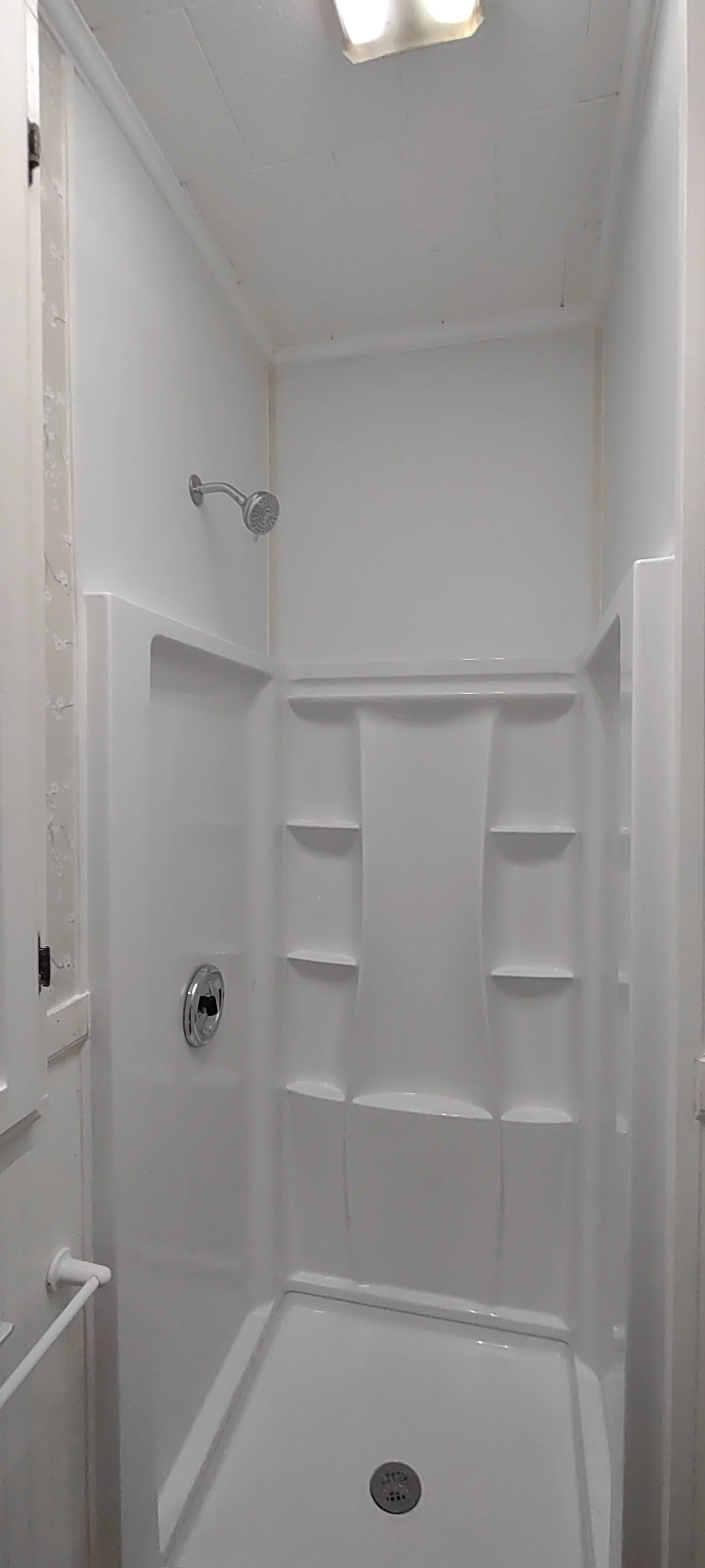 Empty white shower stall with built-in shelves, a showerhead, a drain, and a small window in the ceiling.