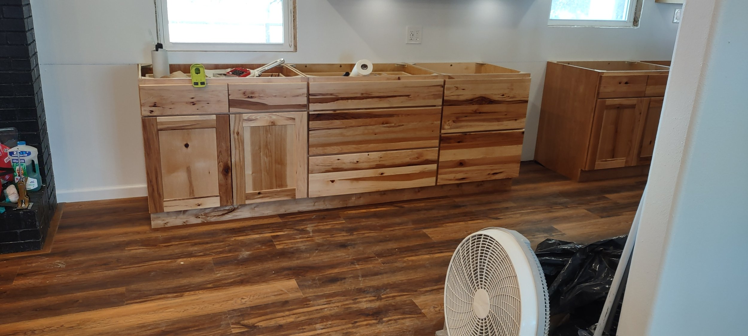 Unfinished wooden kitchen cabinets under construction in a room with hardwood floors and white walls, with tools and supplies on top.