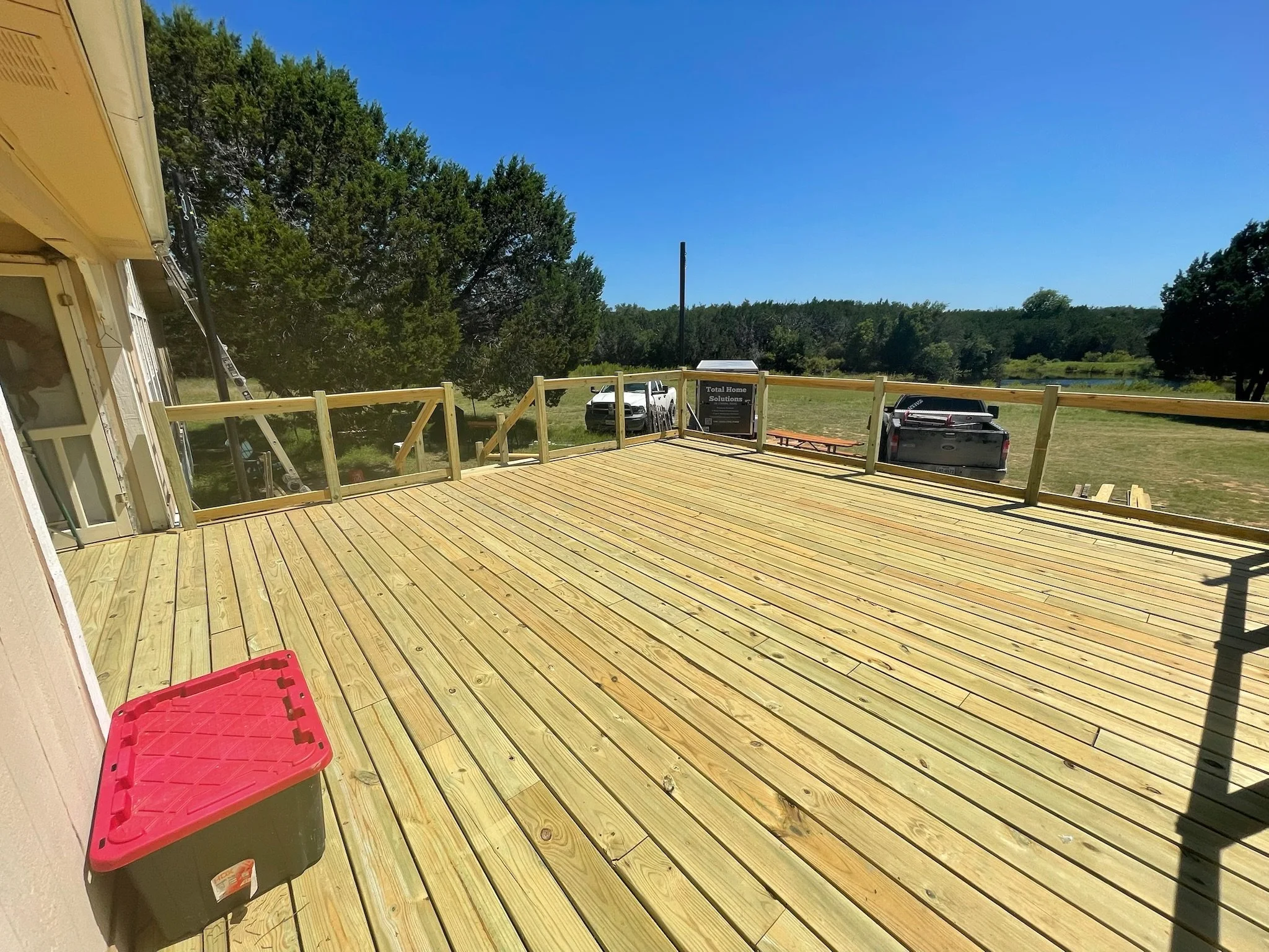 Newly constructed wooden deck outdoors with a red and black storage bin in the foreground, a sign that reads 'Total Home Solutions,' parked trucks, and a view of trees and a river under a clear blue sky.