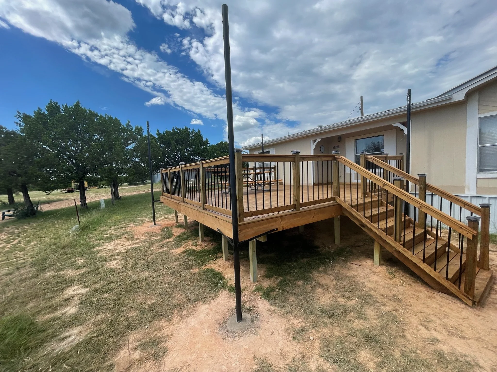 Backyard with wooden deck, black metal railing, set of stairs, and a multi-purpose pole structure, with a one-story house, trees, and a partly cloudy sky in the background.