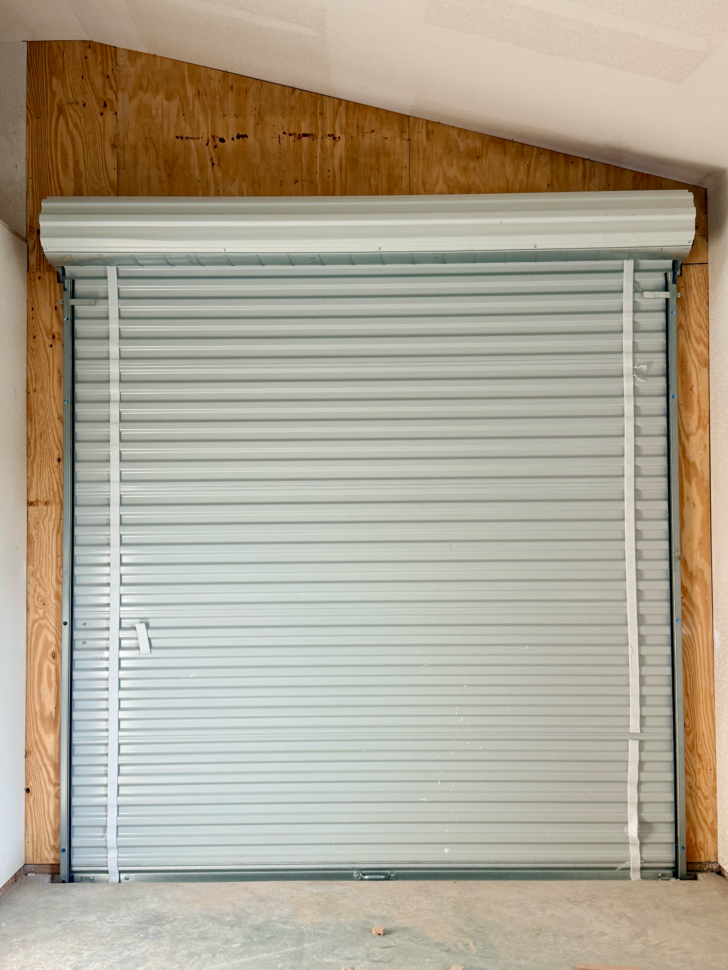 A metal security shutter door installed in a room with unfinished wooden walls and a concrete floor.
