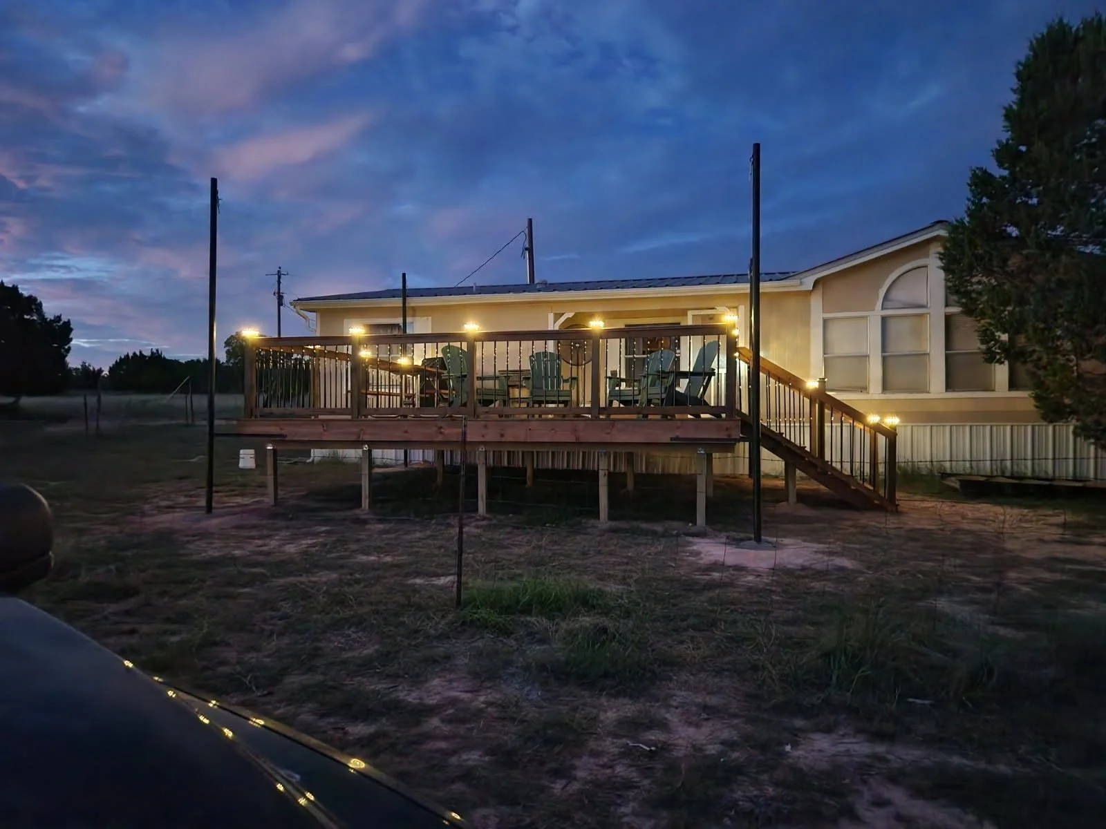 A house with a wooden deck decorated with string lights, outdoor chairs, and stairs leading down to the yard during dusk with a cloudy sky.