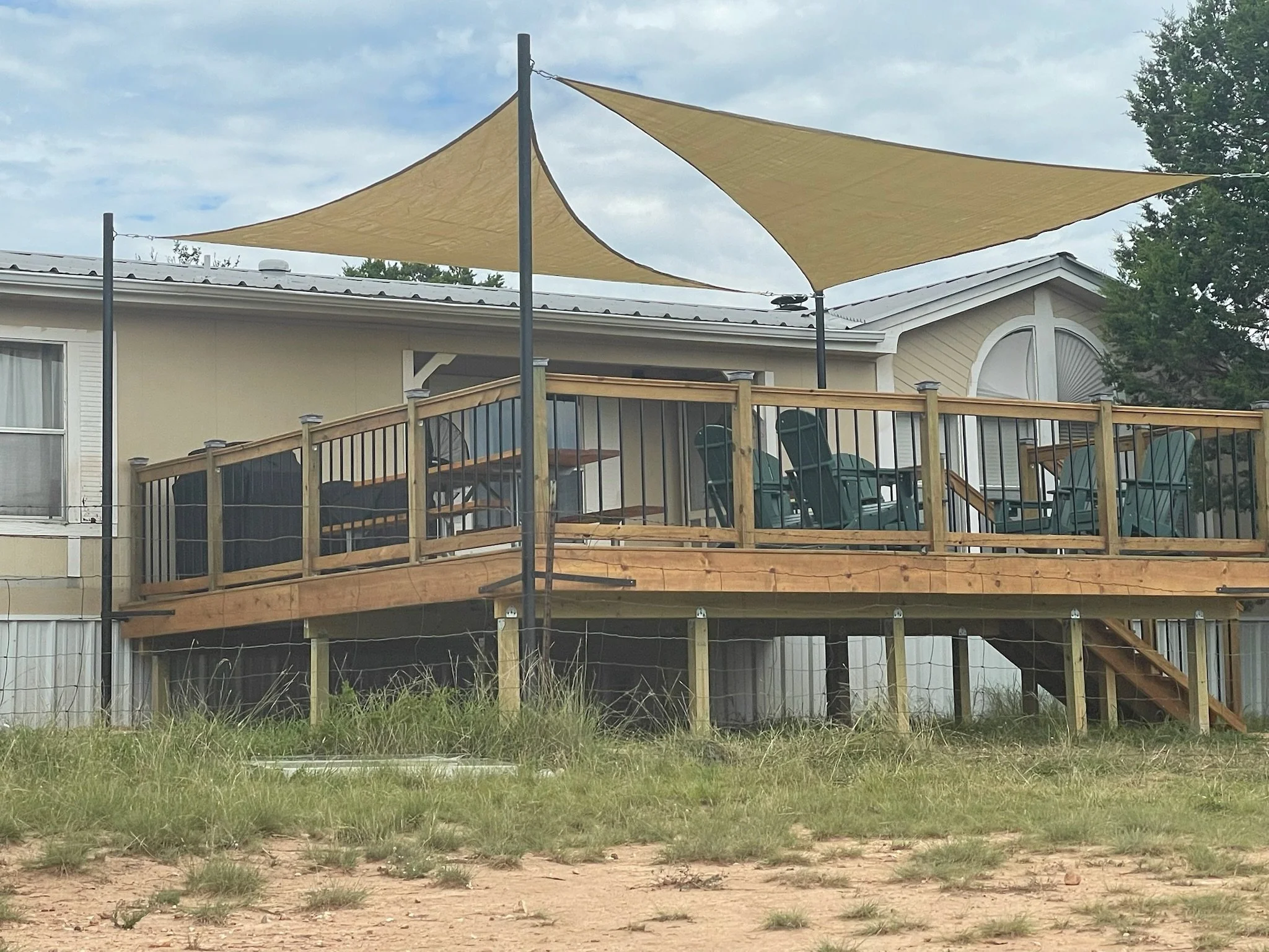 A house with a wooden deck and railing, outdoor chairs, and beige shade sails above the deck, with trees and a cloudy sky in the background.