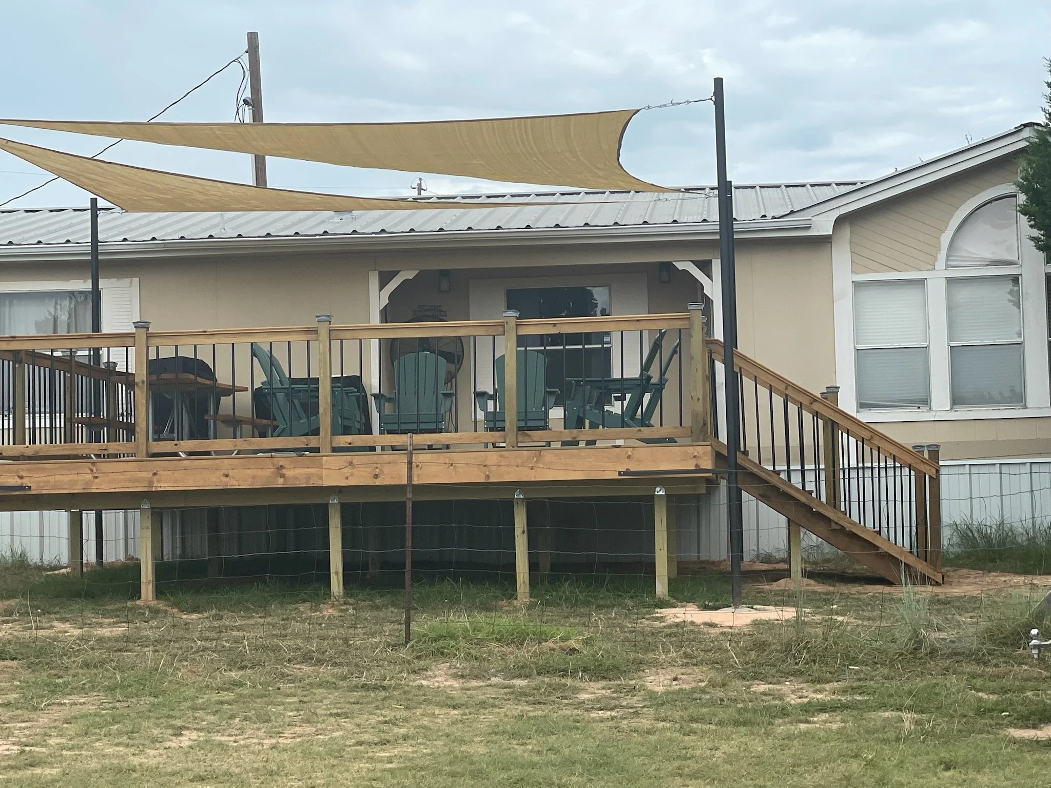 A house with a raised wooden deck and black railing, with outdoor furniture and sail shades overhead, under an overcast sky.