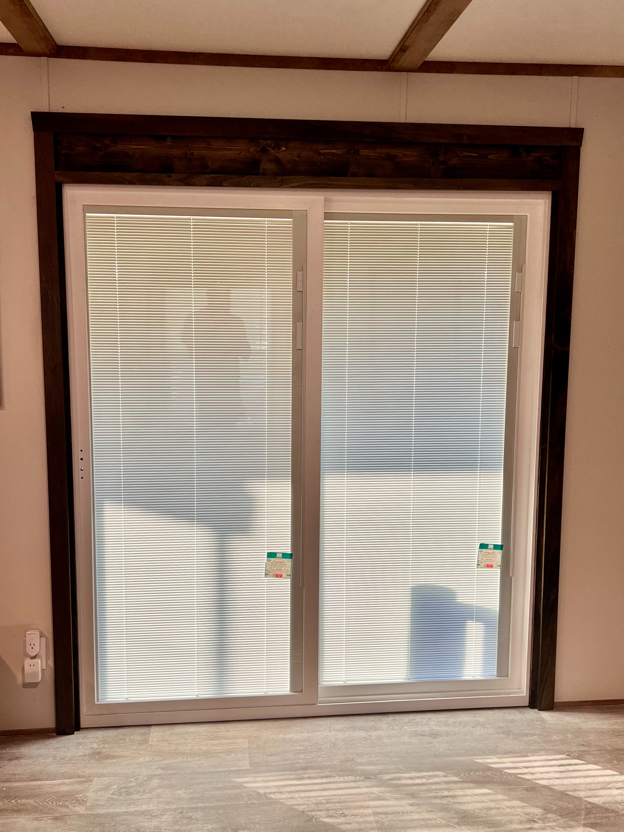 A large glass sliding door with white frame, covered with horizontal white blinds, and surrounded by dark brown wooden trim in a room with light gray wood flooring.