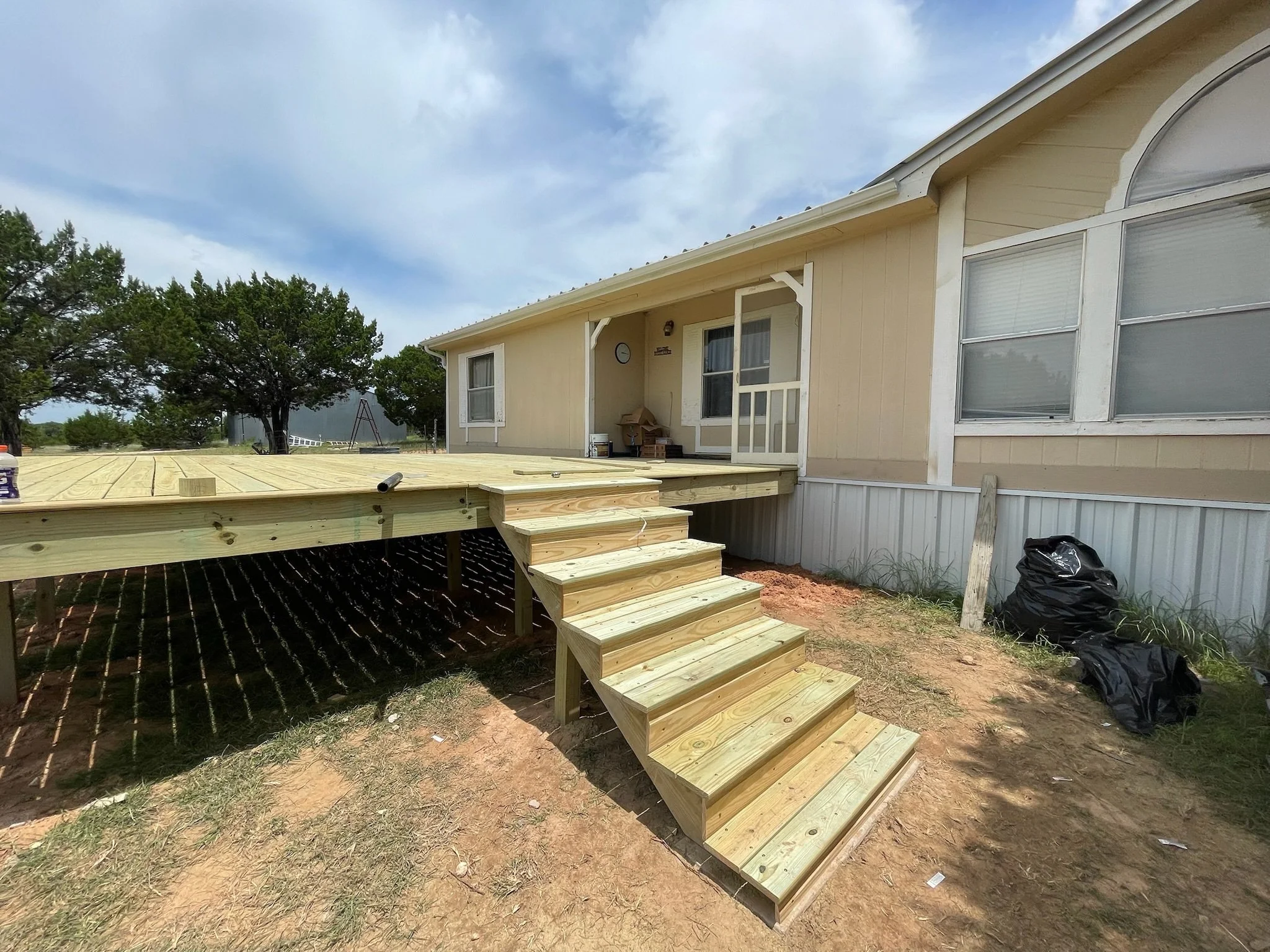 A house exterior with a deck under construction, stairs leading up to the deck, and trees in the background.