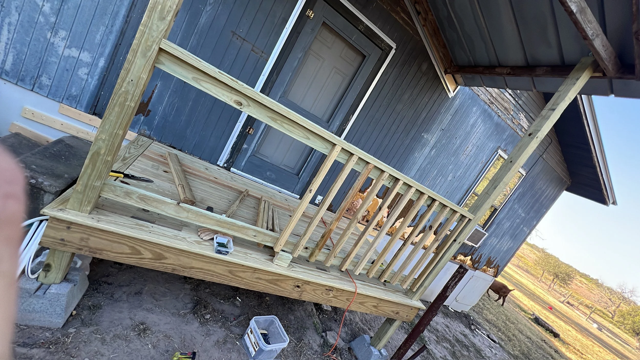 Building a wooden deck with stairs in front of a blue house with a door and window. Construction tools and materials are visible.