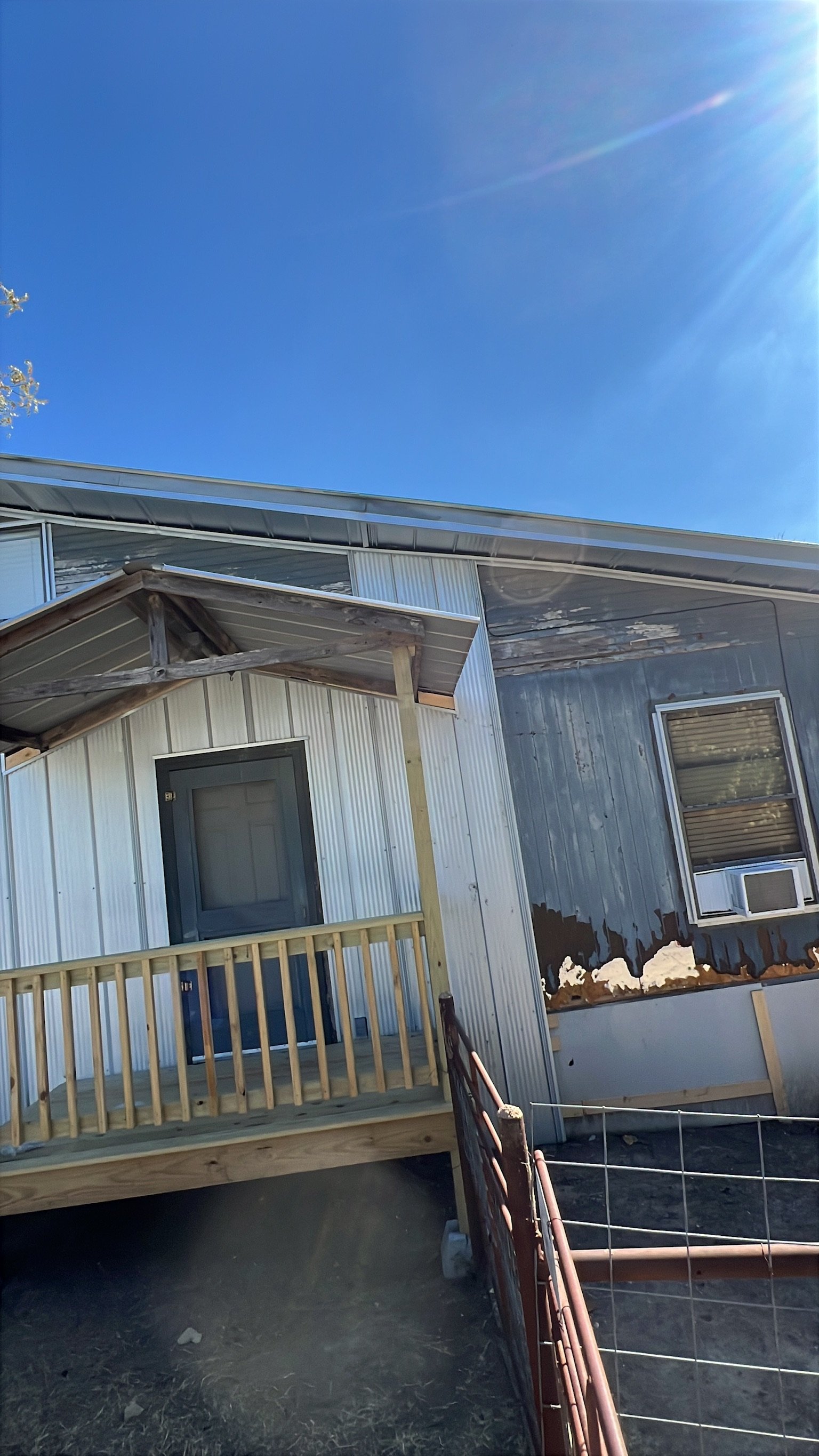 A house with a metal exterior and a small porch under construction, with new railing and support beams. The house has one window with blinds and a window air conditioning unit. The sky is clear and blue with the sun shining brightly.