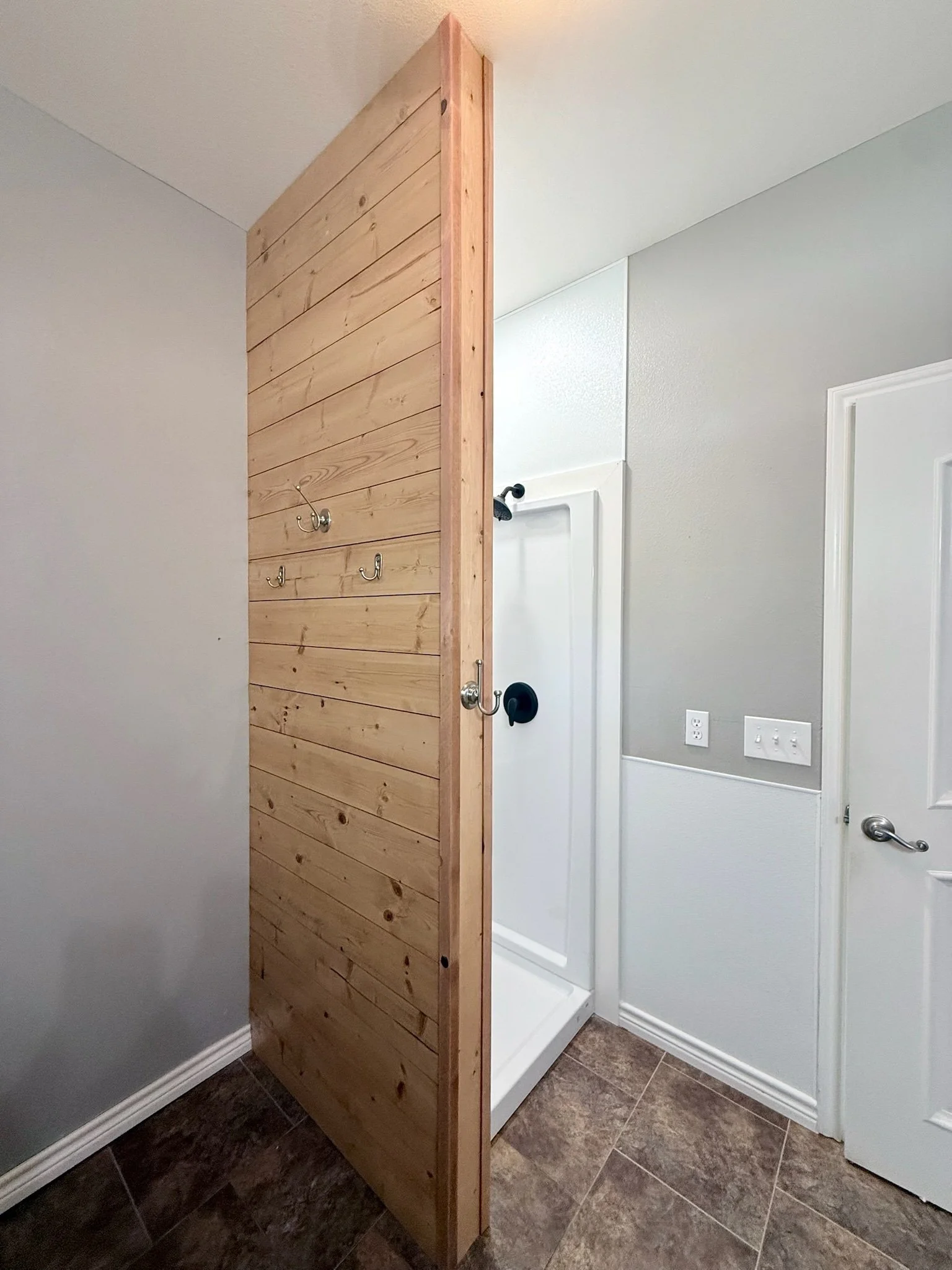 A bathroom with a wooden privacy wall for a shower, with a showerhead visible behind the wall. The bathroom has gray walls, white trim, and a tiled floor.
