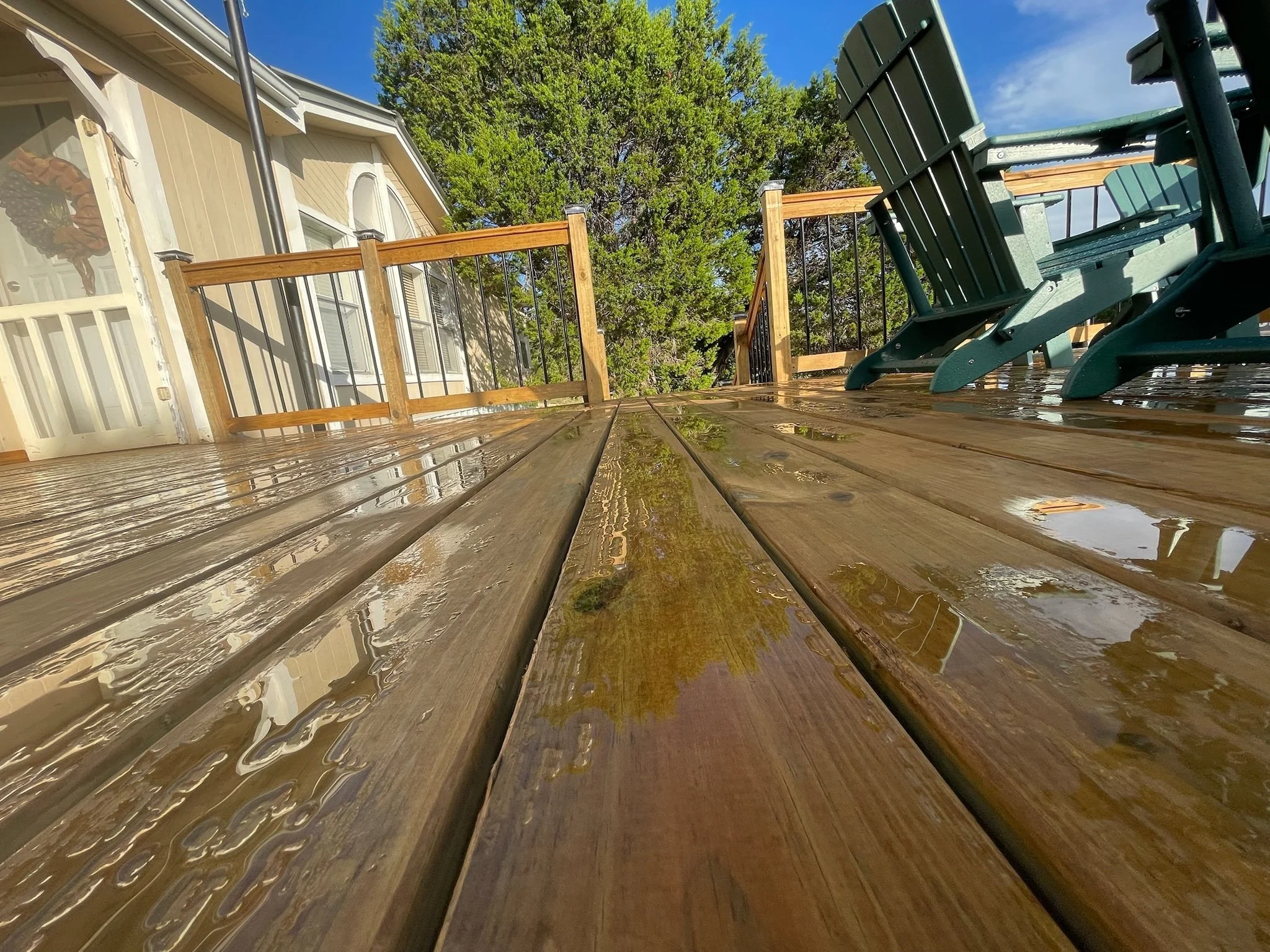 A wooden deck with outdoor furniture including green chairs, a greenery tree in the background, and the house exterior with windows and a door; the sky is blue with some clouds.
