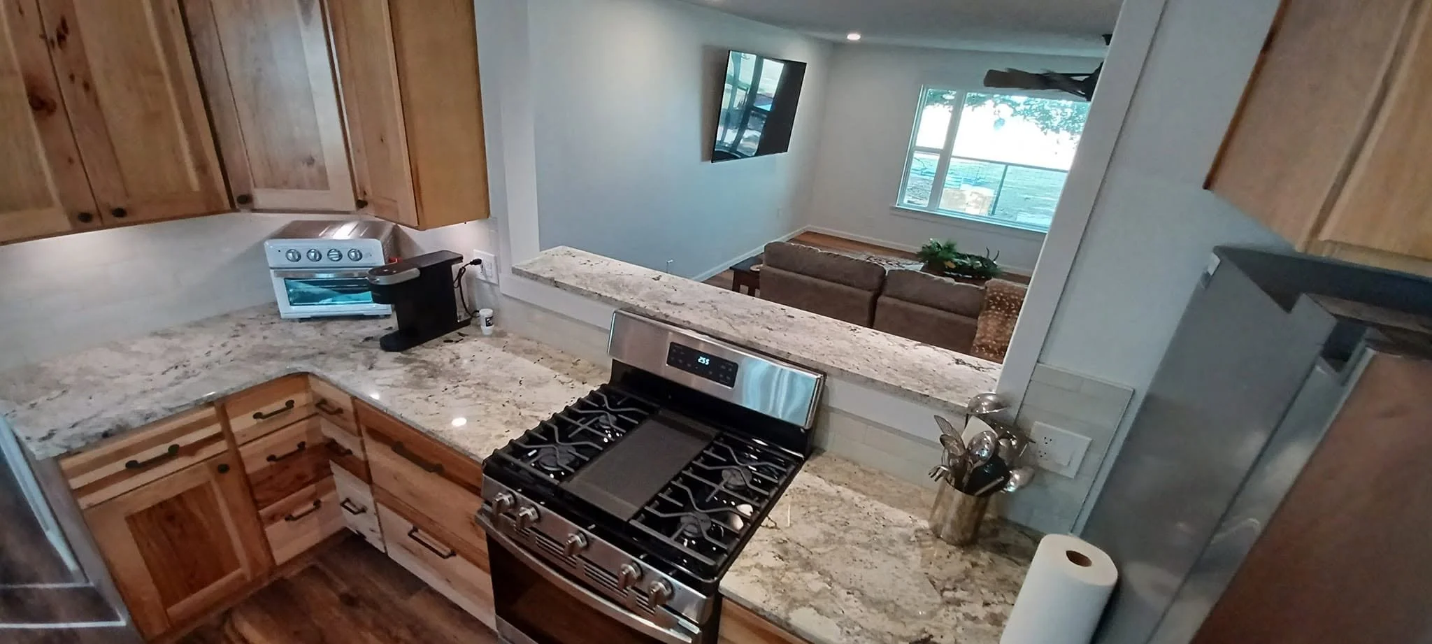Inside view of a kitchen with wooden cabinets, a granite countertop, a stainless steel stove, and a partial view of a living room with a couch and window