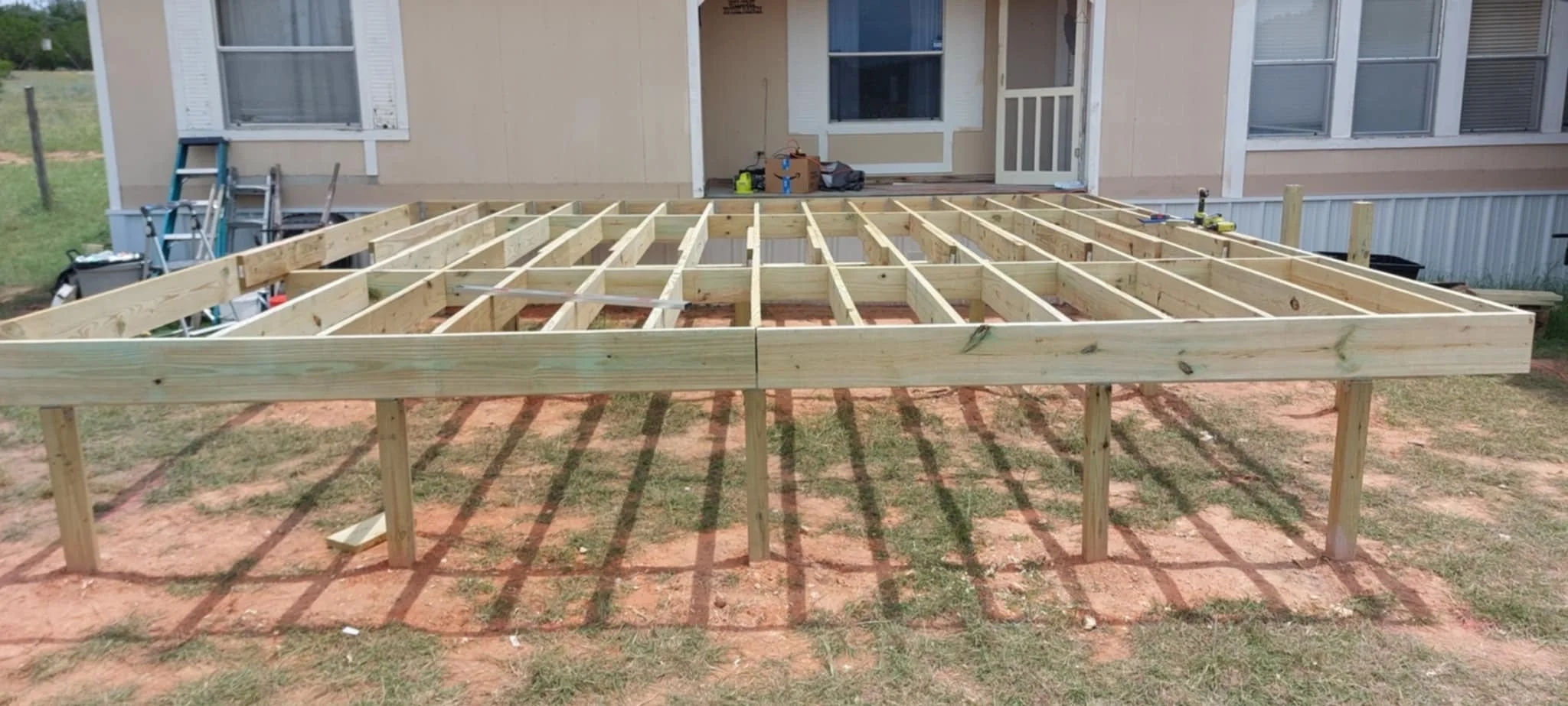 Construction site with a wooden deck frame in front of a house, tools and construction materials nearby.