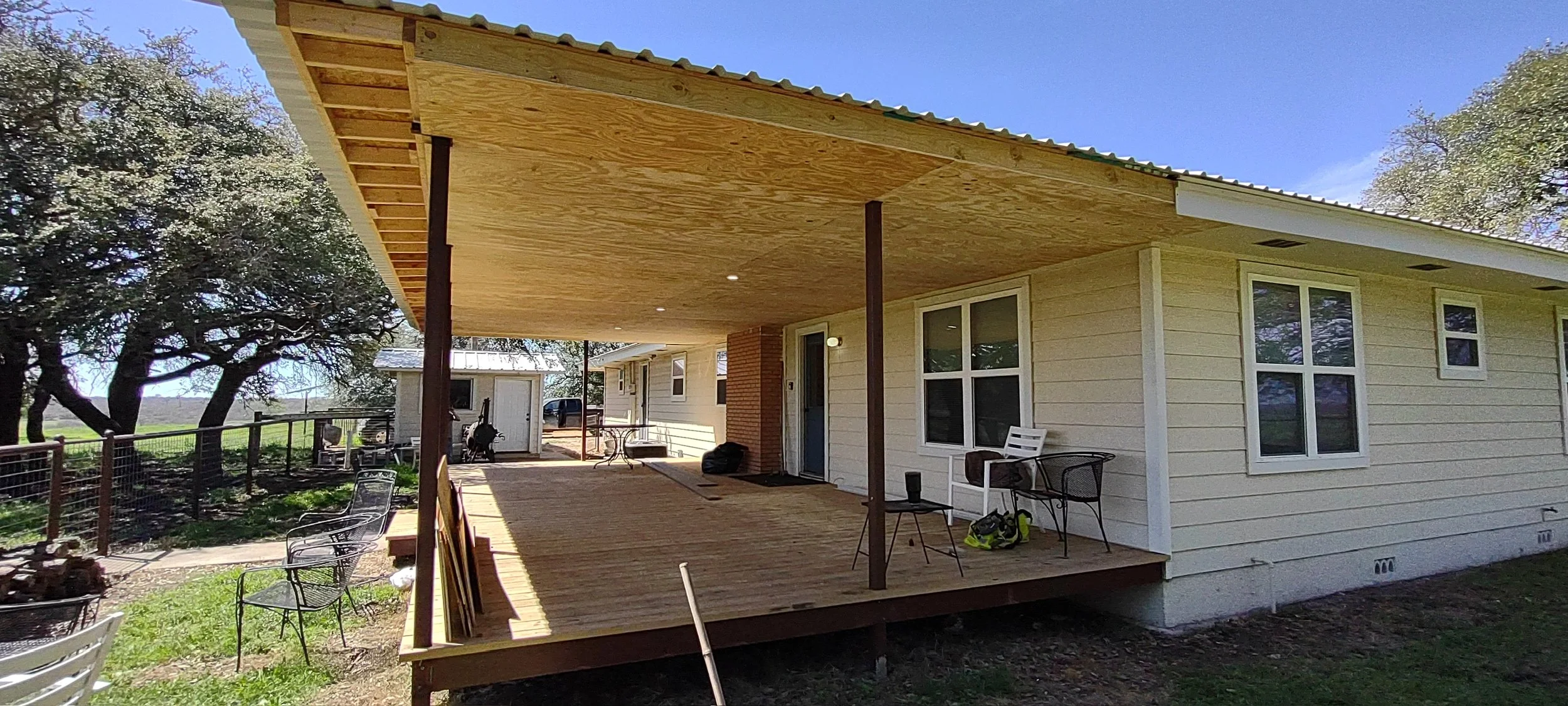 Resized house with a spacious wooden porch, supported by metal posts, with outdoor furniture and tools, set in a rural area with trees and open landscape.
