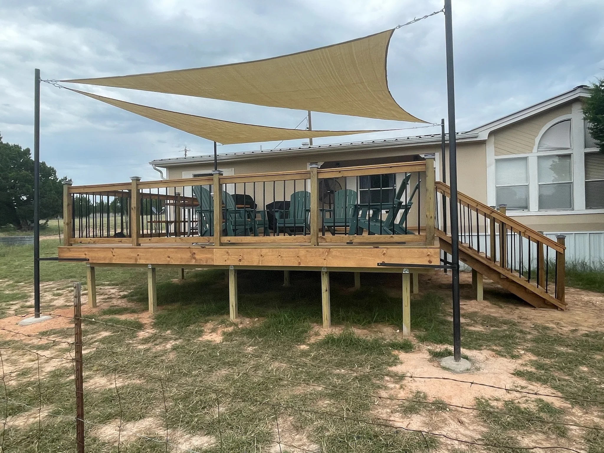 A backyard wooden deck with a beige shade sail overhead, chairs underneath, railing around, and a house with a window and sliding door in the background. There is a fence and open outdoor space nearby.