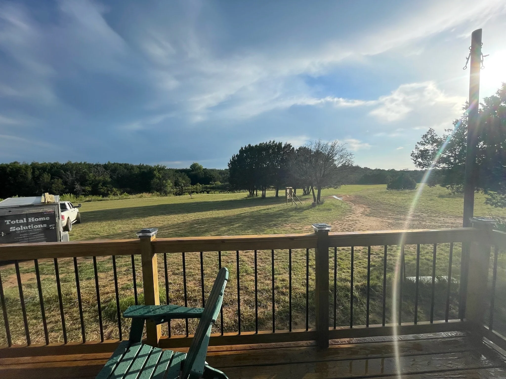 View through a window showing a grassy yard with trees, a driveway, a white truck with text on it, and a blue sky with scattered clouds. Sunlight is shining through the window.