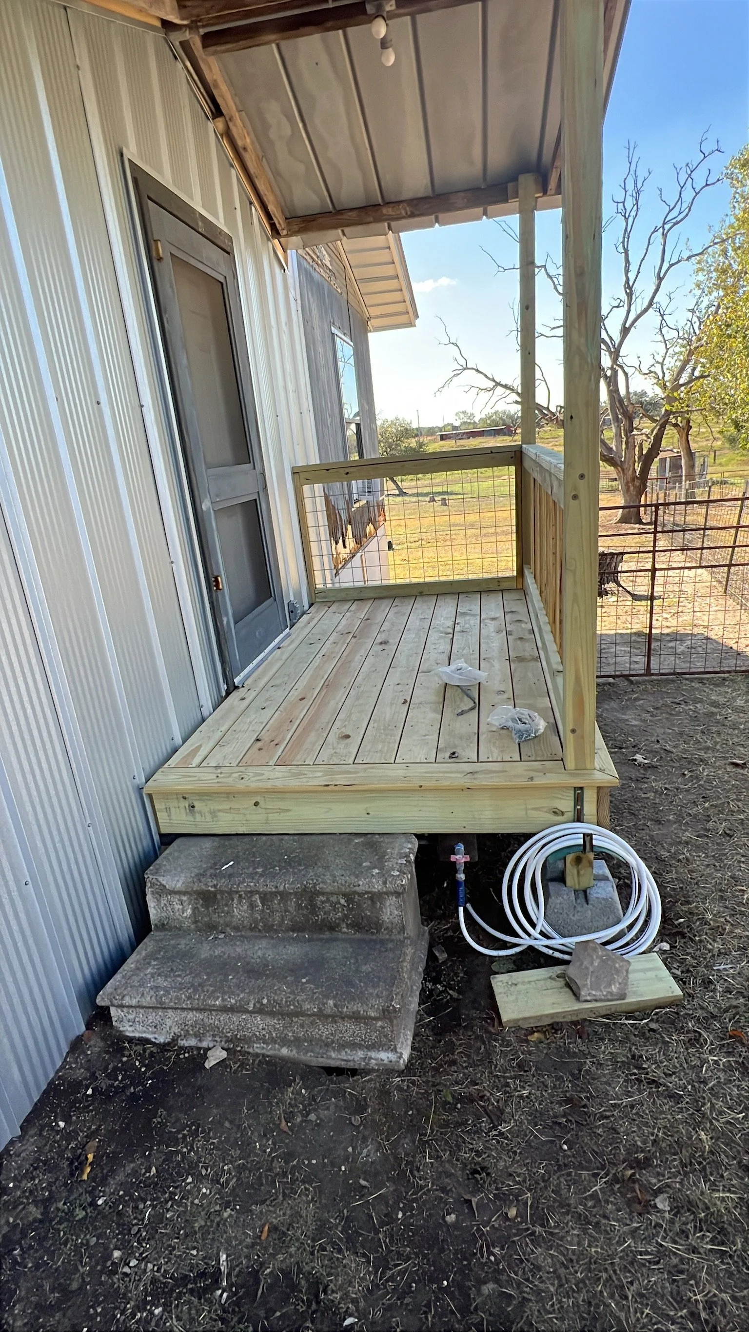 A small wooden porch with steps leading up to it, attached to the side of a metal building. The porch has a railing on right side, and construction materials including coiled tubing and a small piece of wood nearby. In the background, there are trees
