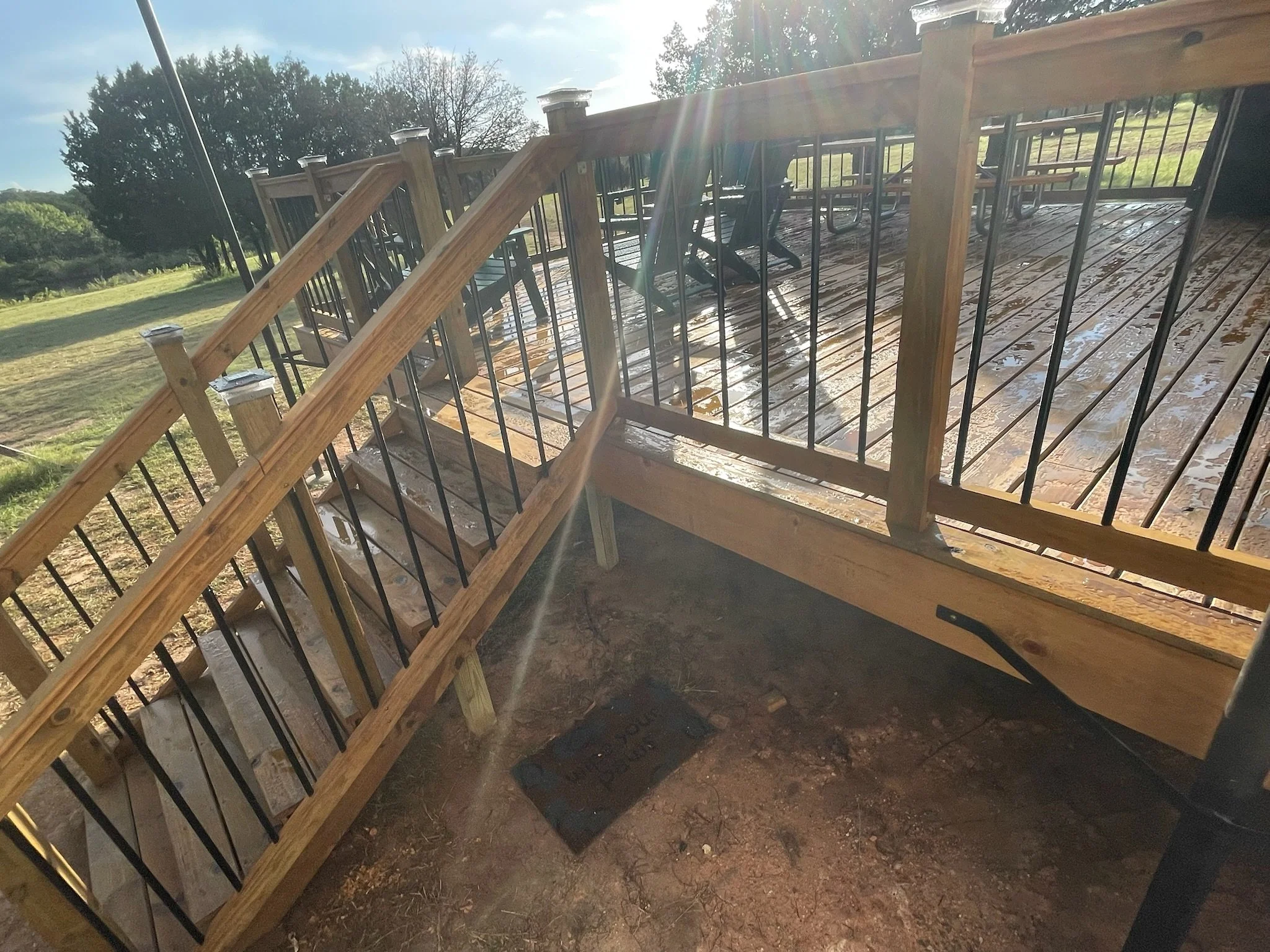 Newly installed wooden and metal staircase leading up to a wooden deck outdoors, with green trees and a grassy field in the background, sunny weather with some clouds.