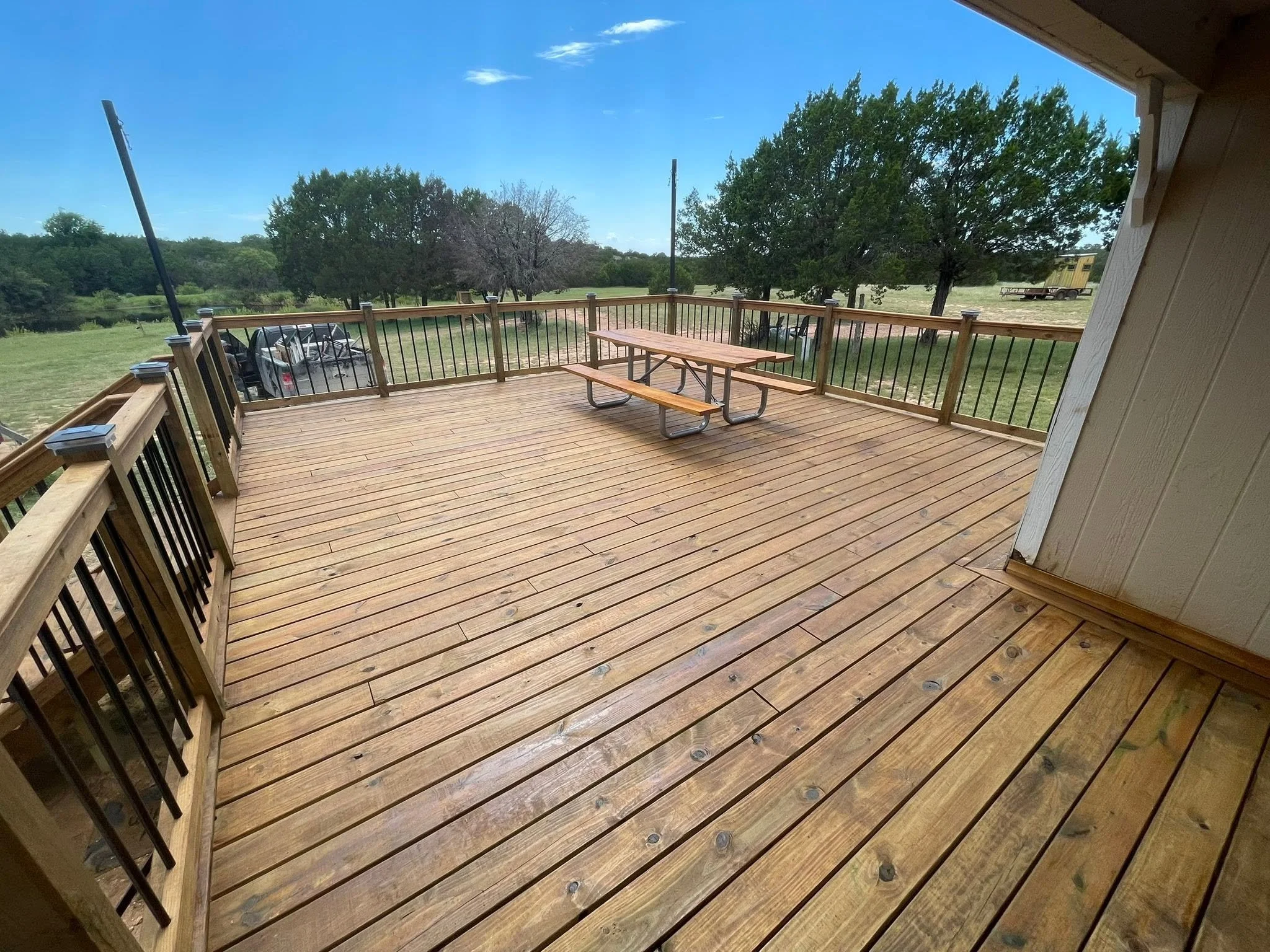 Wooden deck with picnic table, black metal railing, trees, grassy field, car, utility pole, and a yellow building or trailer in the background under a blue sky with some clouds.
