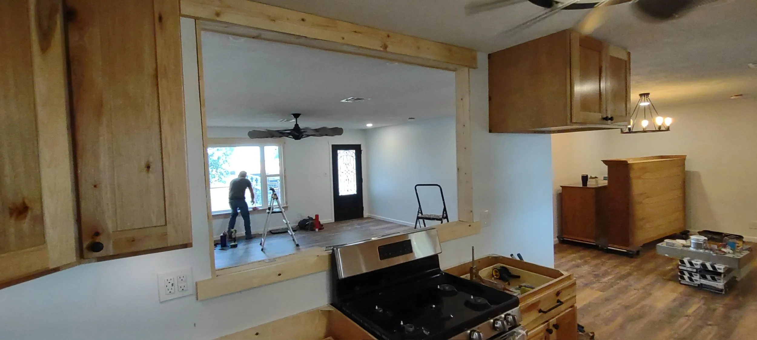Interior of a house under renovation with a man working near a large window, ladder, wooden cabinets, and construction tools scattered around.