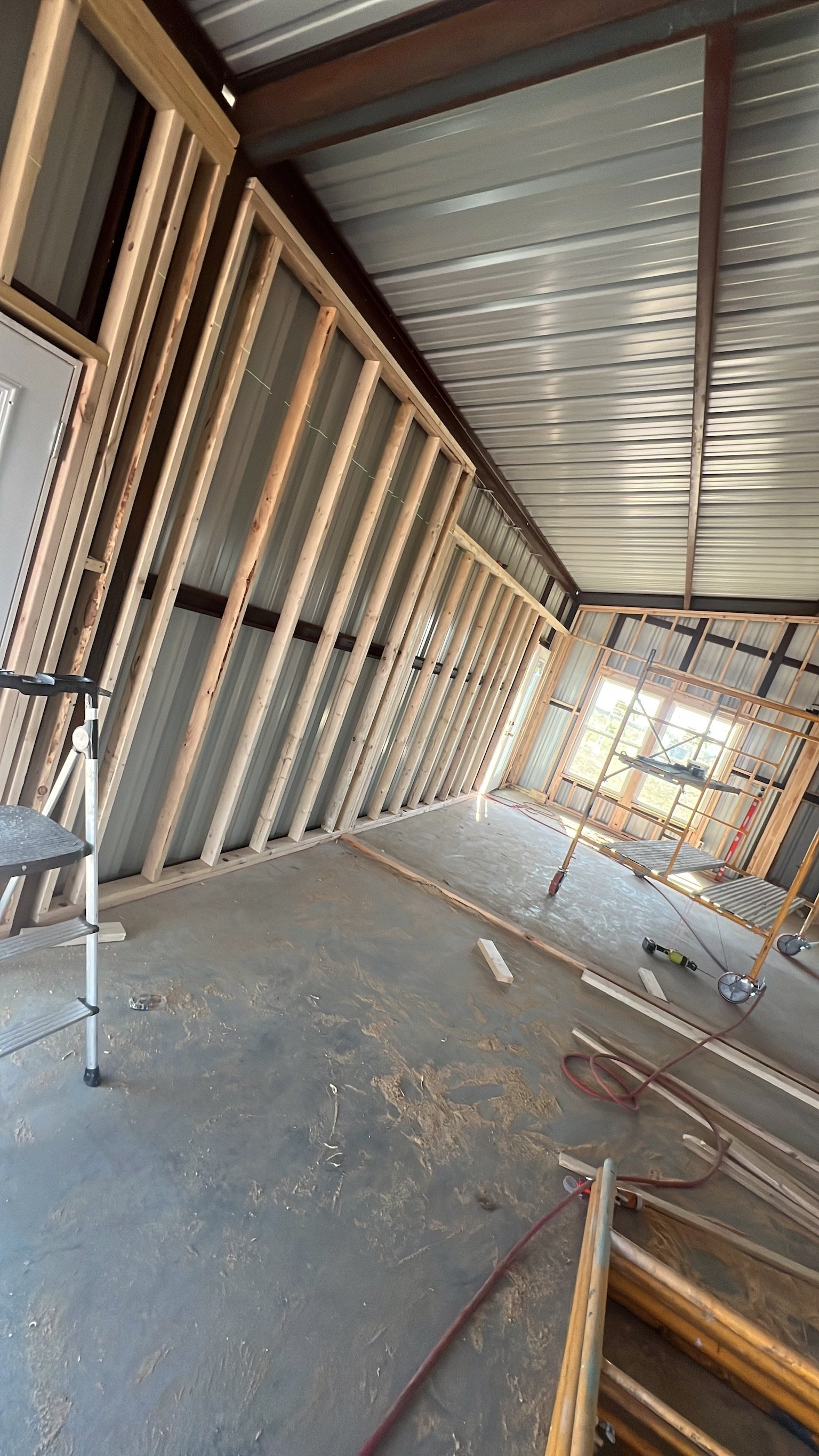 Interior construction site with wooden framing, metal wall panels, scaffolding, and construction tools on a concrete floor.