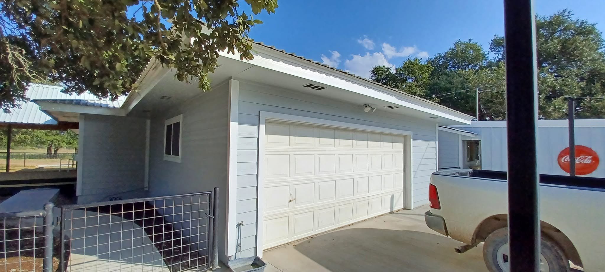 A residential house with a white garage door and a white exterior, surrounded by a metal fence and a white truck parked nearby, on a sunny day with green trees and a blue sky.