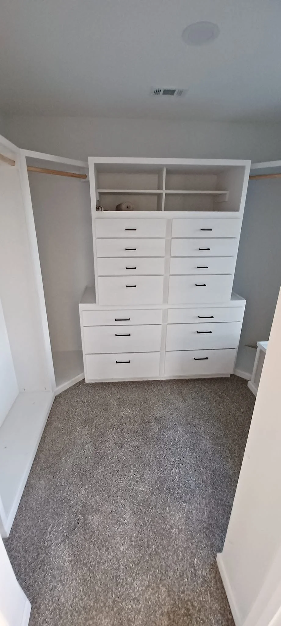 Empty closet with white built-in drawers and shelves and beige carpeting.