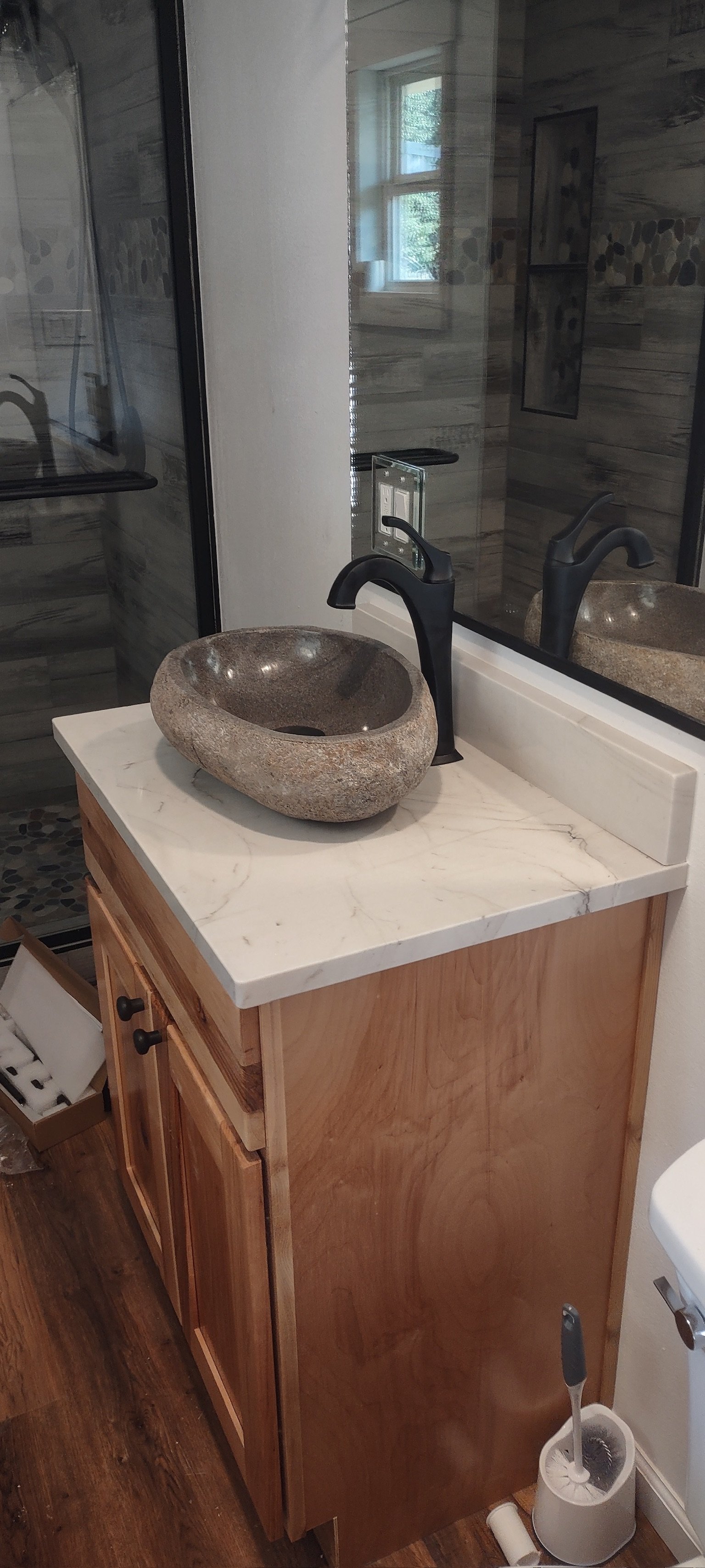 A bathroom vanity with a stone vessel sink, black faucet, and marble countertop. Behind it, a large mirror reflects a window and part of a shower area with gray tile walls.