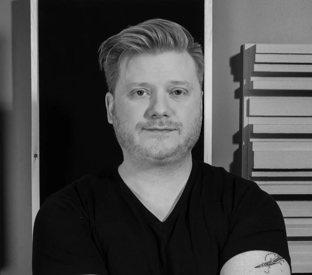 Black and white portrait of a young man with styled hair and a beard, crossing his arms, standing indoors near a wall and acoustic foam panels.