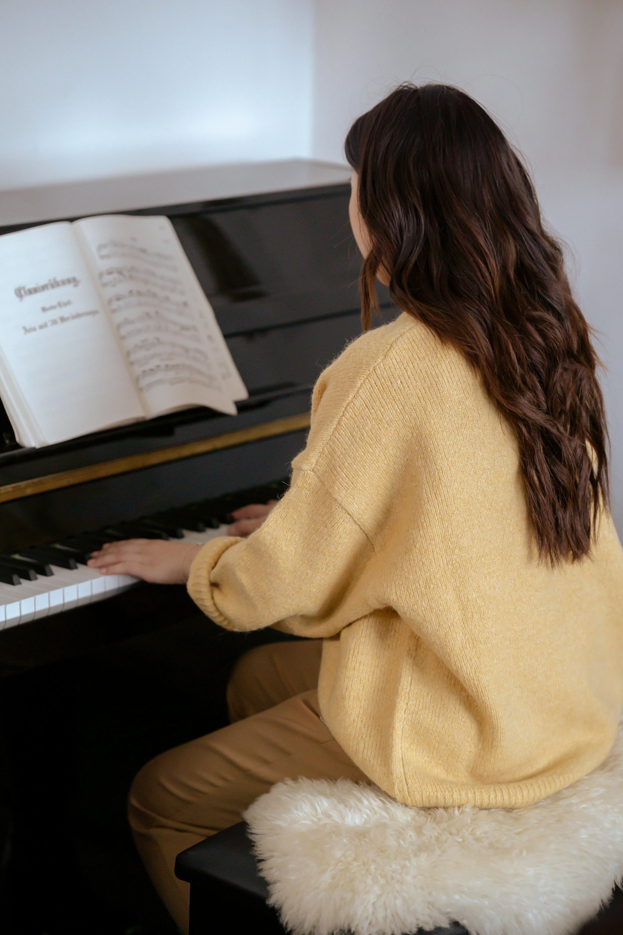 A person with long brown wavy hair wearing a yellow sweater, sitting on a cushioned stool, playing the piano.