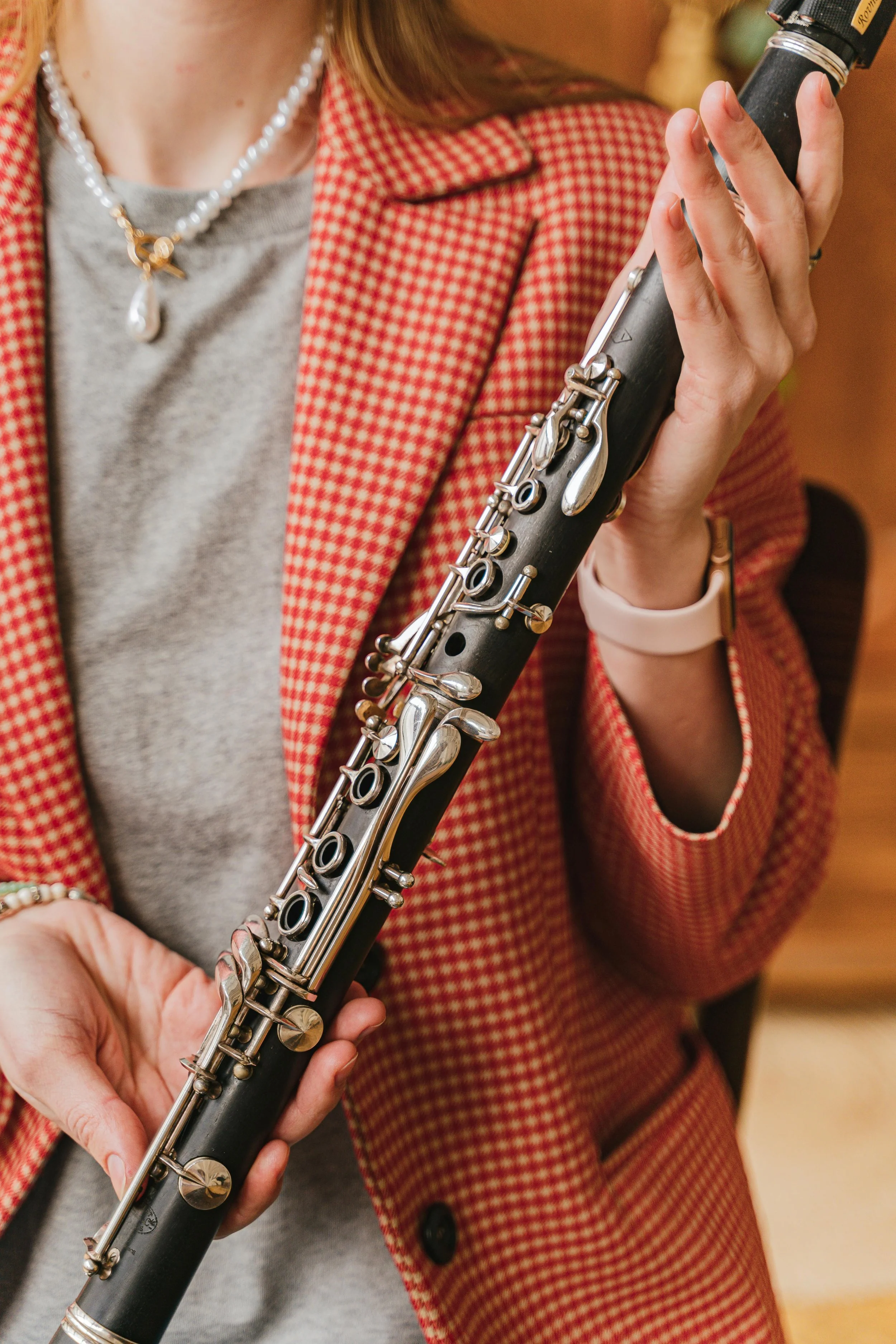 Close-up of a person holding a clarinet, wearing a red checkered blazer, a grey shirt, and a pearl necklace.