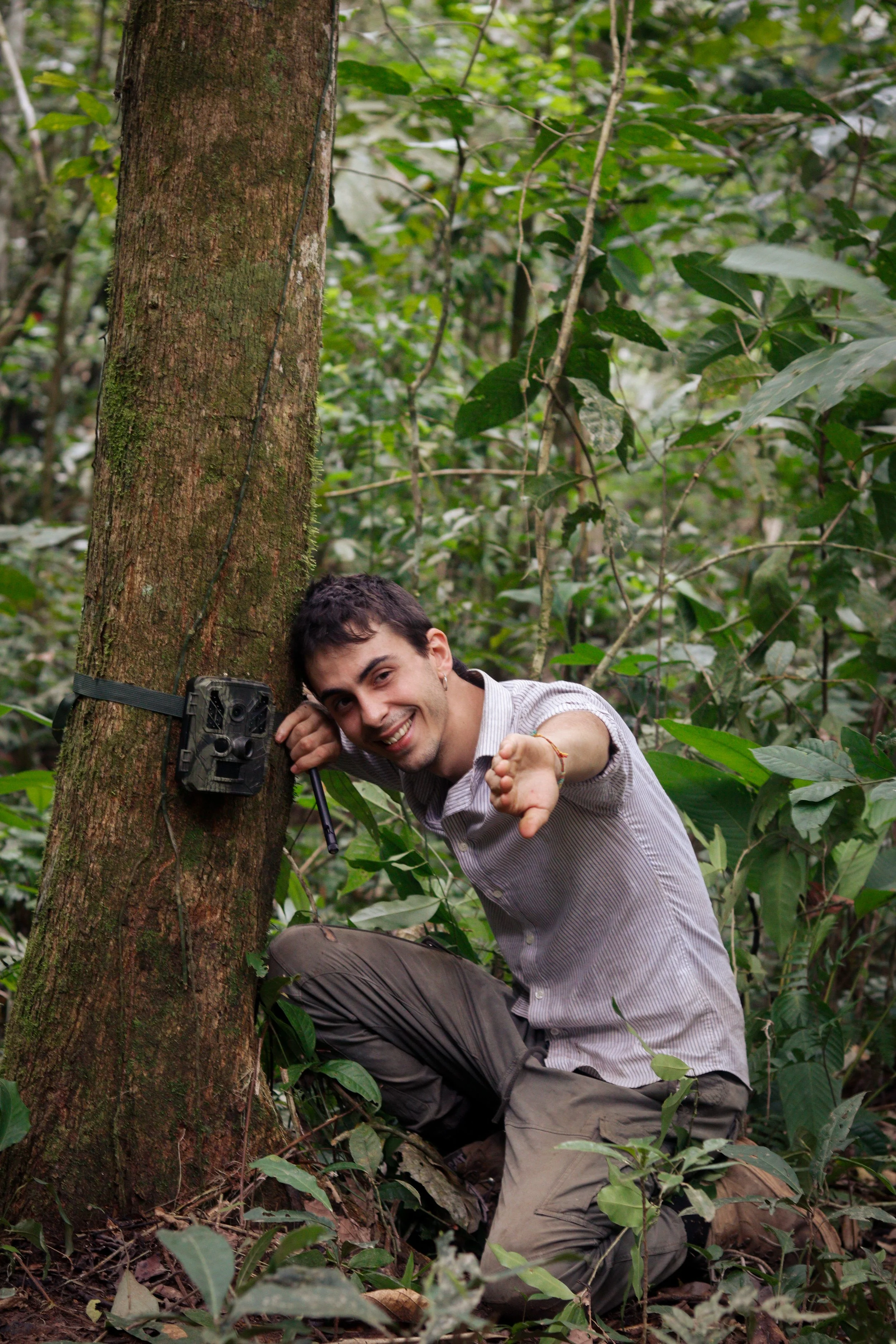 A young man in a forest kneeling next to a tree, smiling and reaching out with his hand, with a trail camera attached to the tree.