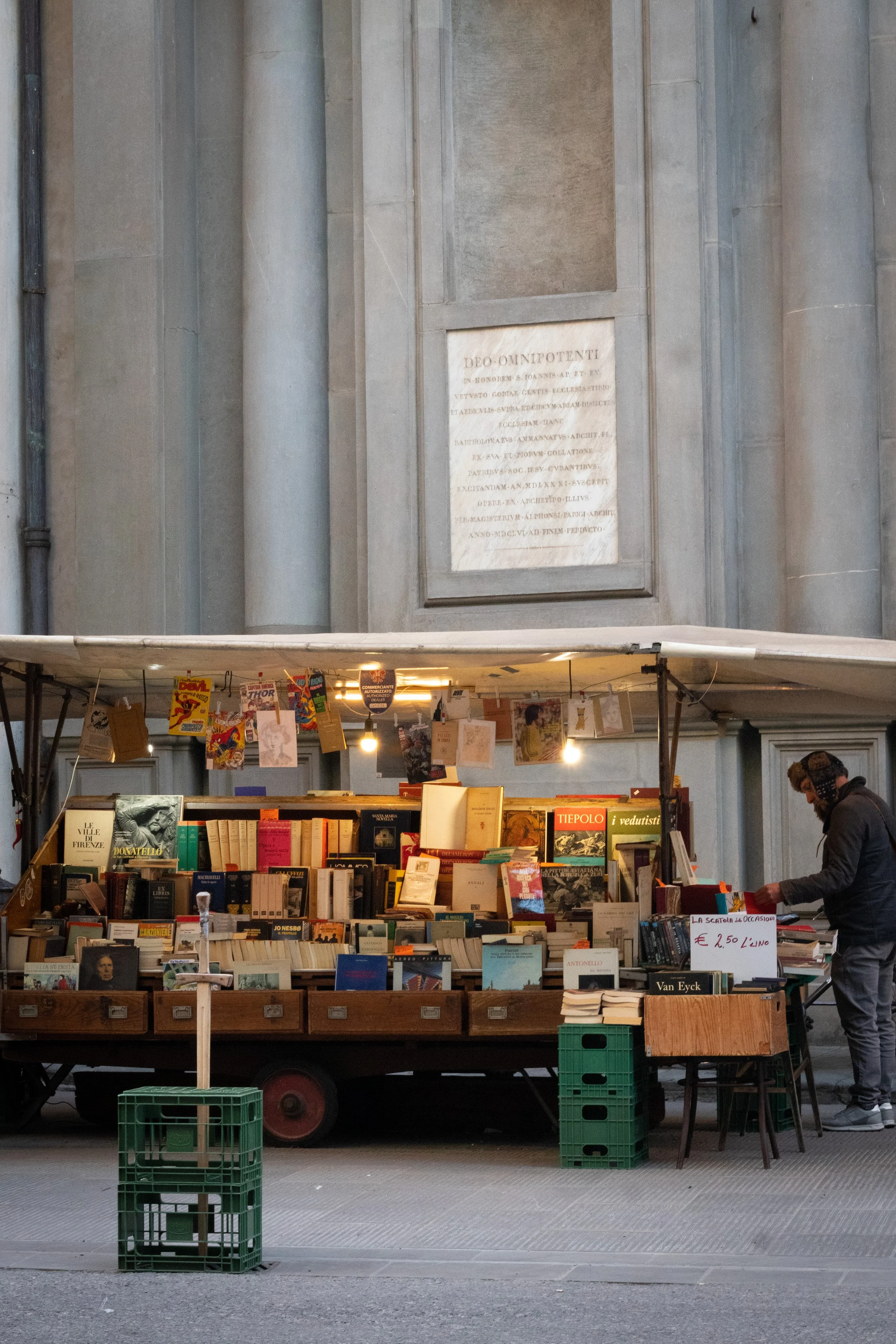 A street book stand with various books and postcards, set against a historical building with a large plaque on the wall.