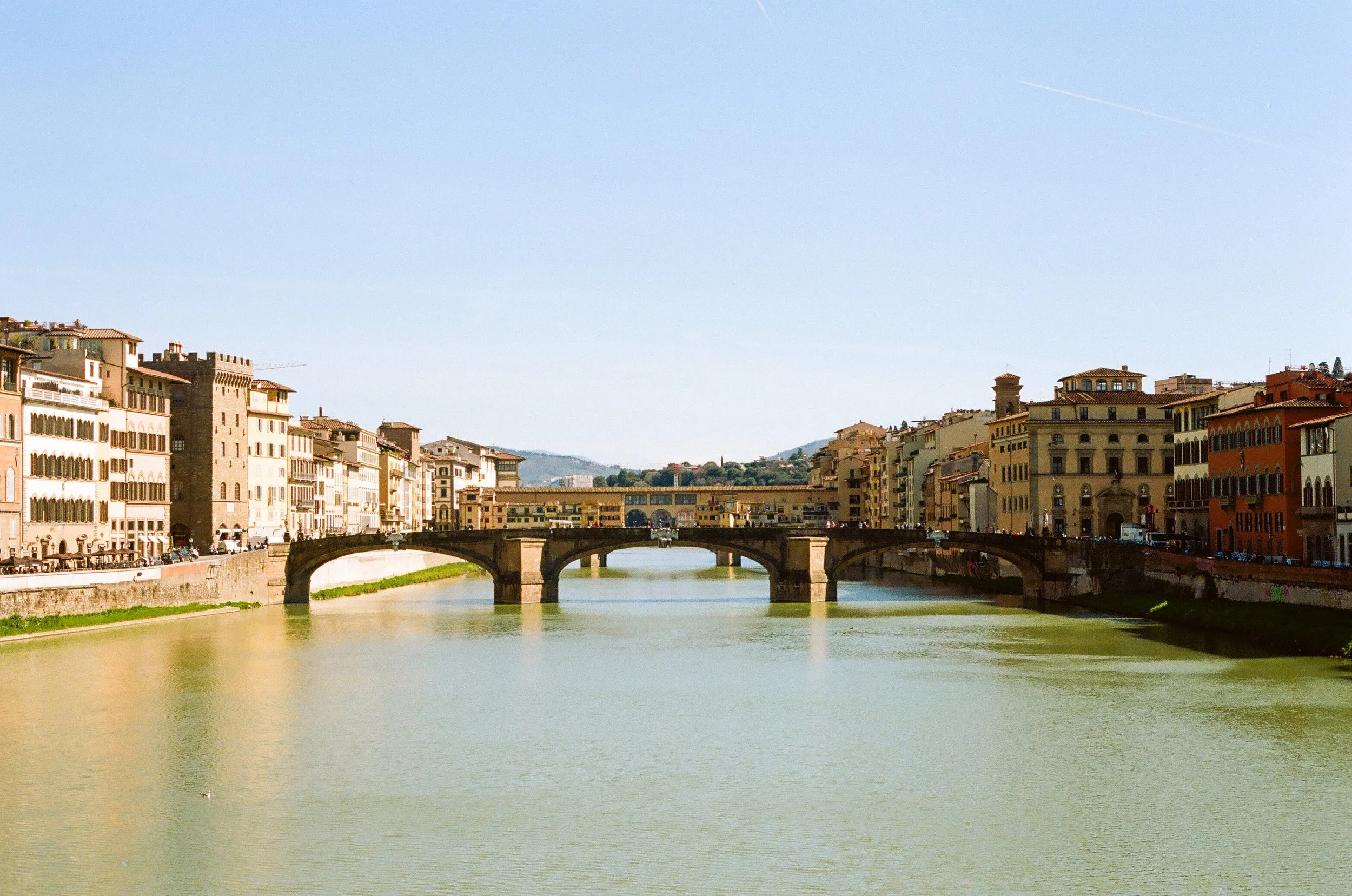 A scenic view of a river with a stone bridge and historic buildings lining both sides, under a clear blue sky.