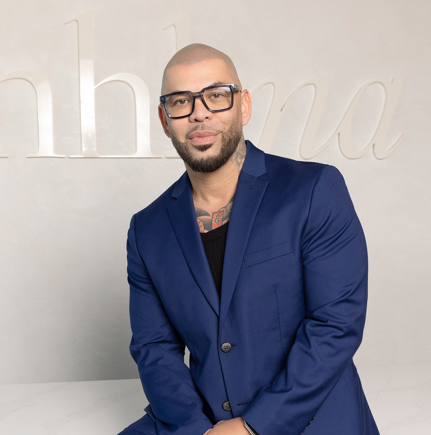 A man wearing a blue suit and black-rimmed glasses poses in front of a light-colored wall with blurred text in the background.