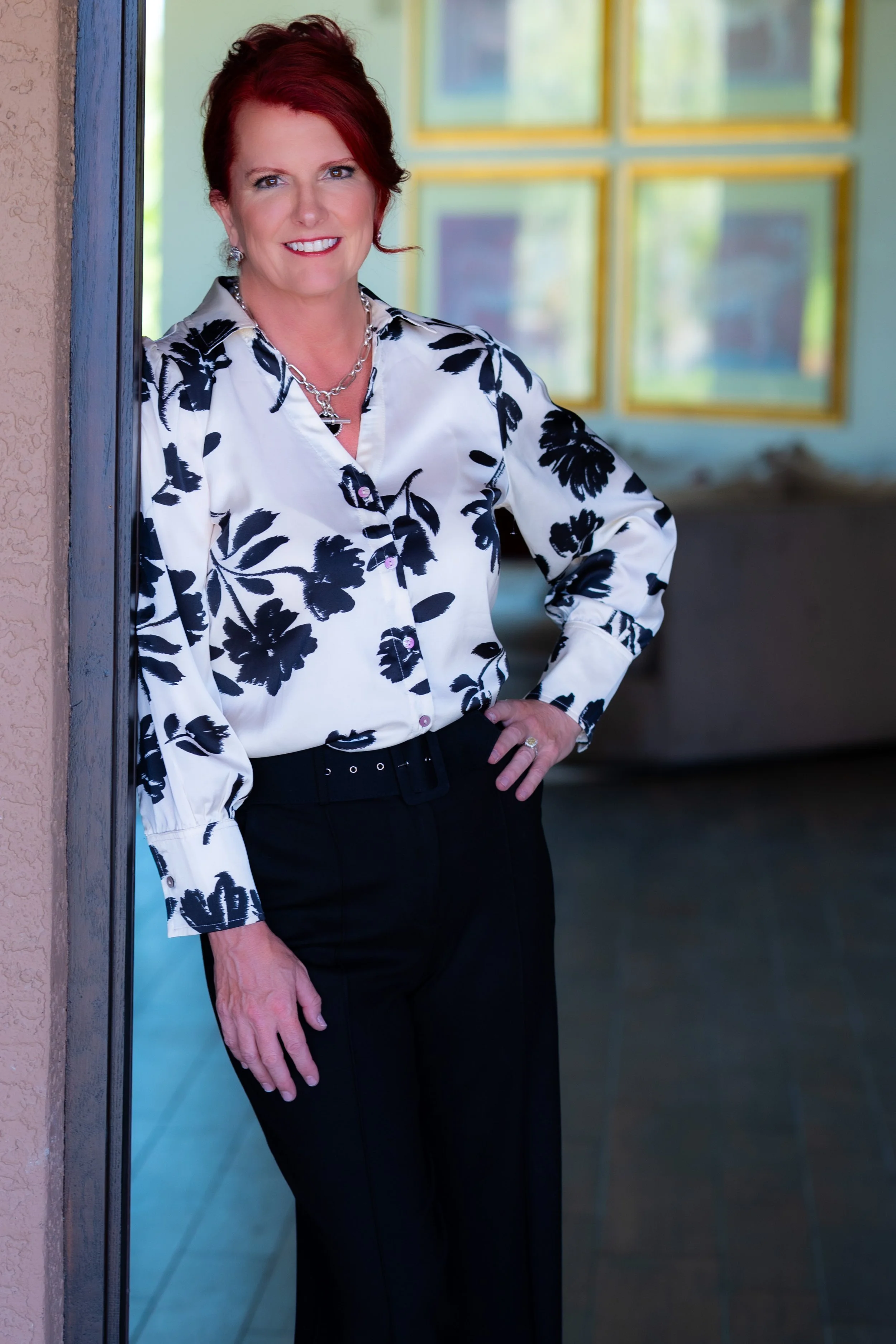 A woman with short red hair, wearing a white blouse with black floral pattern and black pants, standing indoors near a textured wall and colorful windows, smiling at the camera.