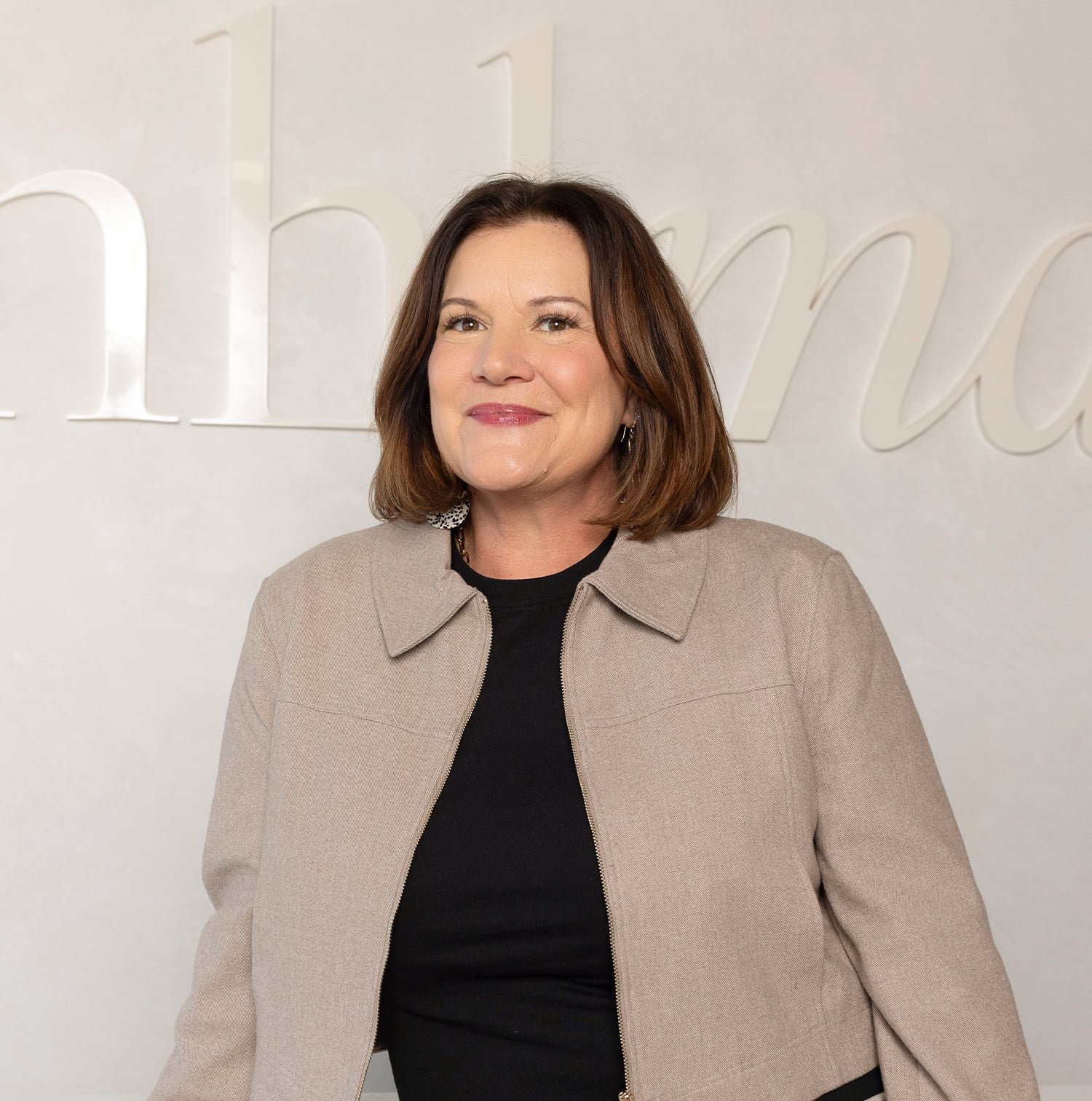 Woman with shoulder-length brown hair wearing a beige jacket and black top standing in front of a wall with partially visible stylized text.